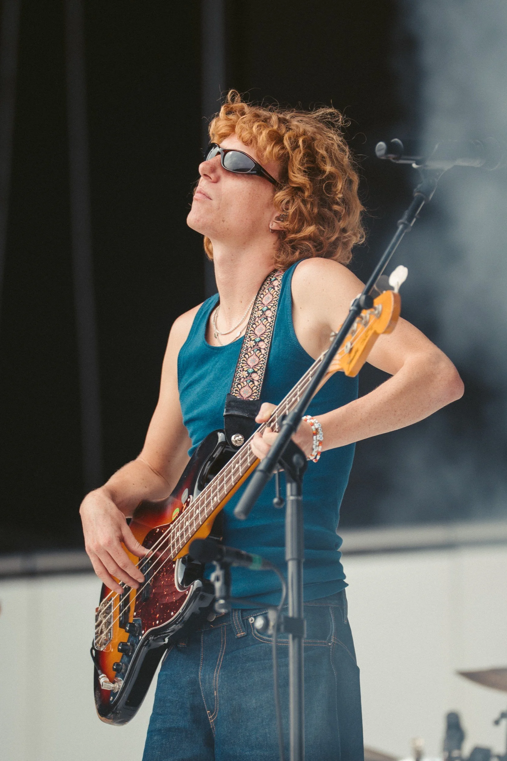 Junge Frau mit roten Locken, Sonnenbrille, blauer Tanktop und Armbändern spielt E-Bass auf einer Bühne.