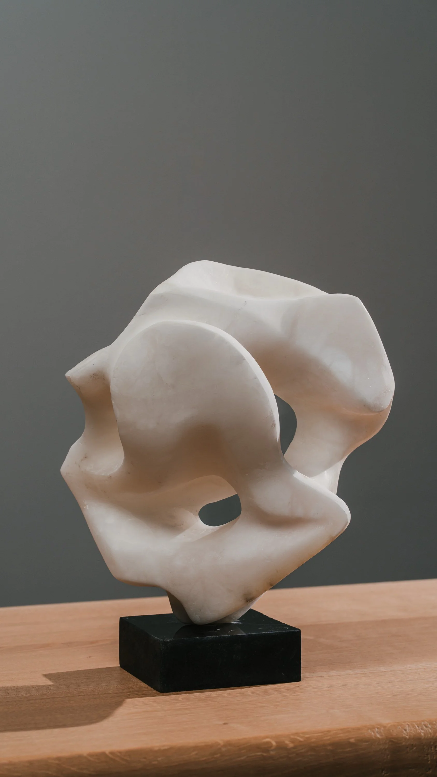 Studio-Fotografie von einem Kunstwerk. Abstrakte weiße Skulptur auf Holzsockel vor grauem Hintergrund.