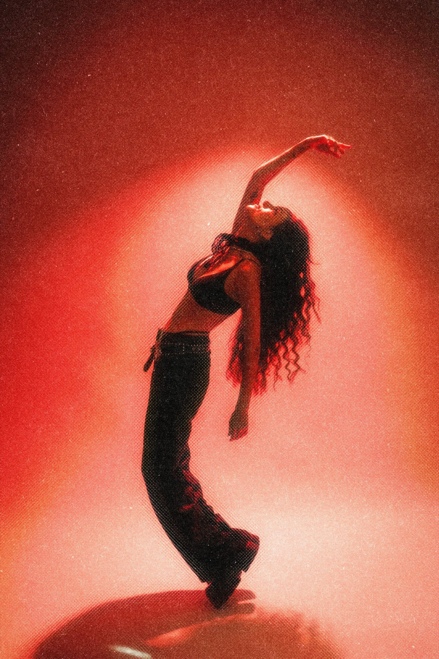 Eine Frau tanzt in einem dunklen Raum mit rotem Licht, trägt schwarze Kleidung, hebt den Arm und bewegt sich im Tanz.