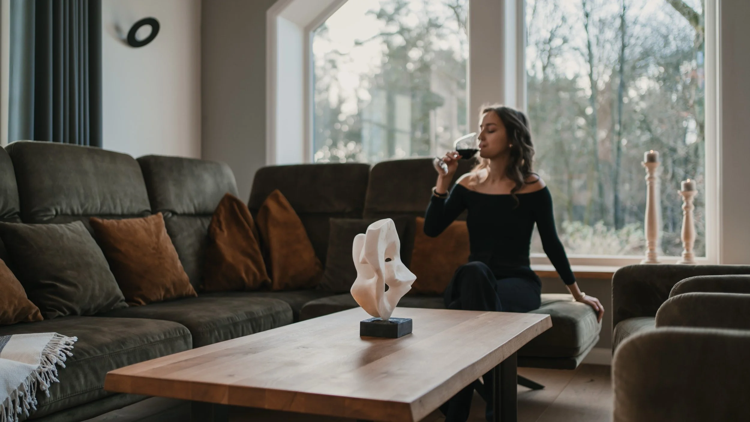 Frau trinkt Rotwein auf einer Couch im Wohnzimmer mit dunklen Sofakissen und einem Holztisch, auf dem eine Skulptur steht, vor einem großen Fenster mit Blick ins Freie.