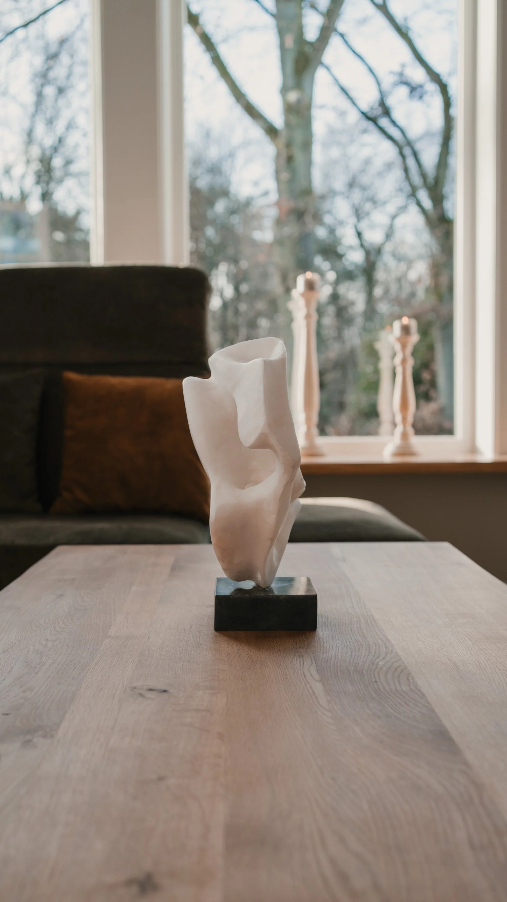 Kunstvolle weiße Skulptur auf einem Holztablett vor einem Fenster mit Blick auf Bäume, im Hintergrund Kerzen auf Fensterbank.