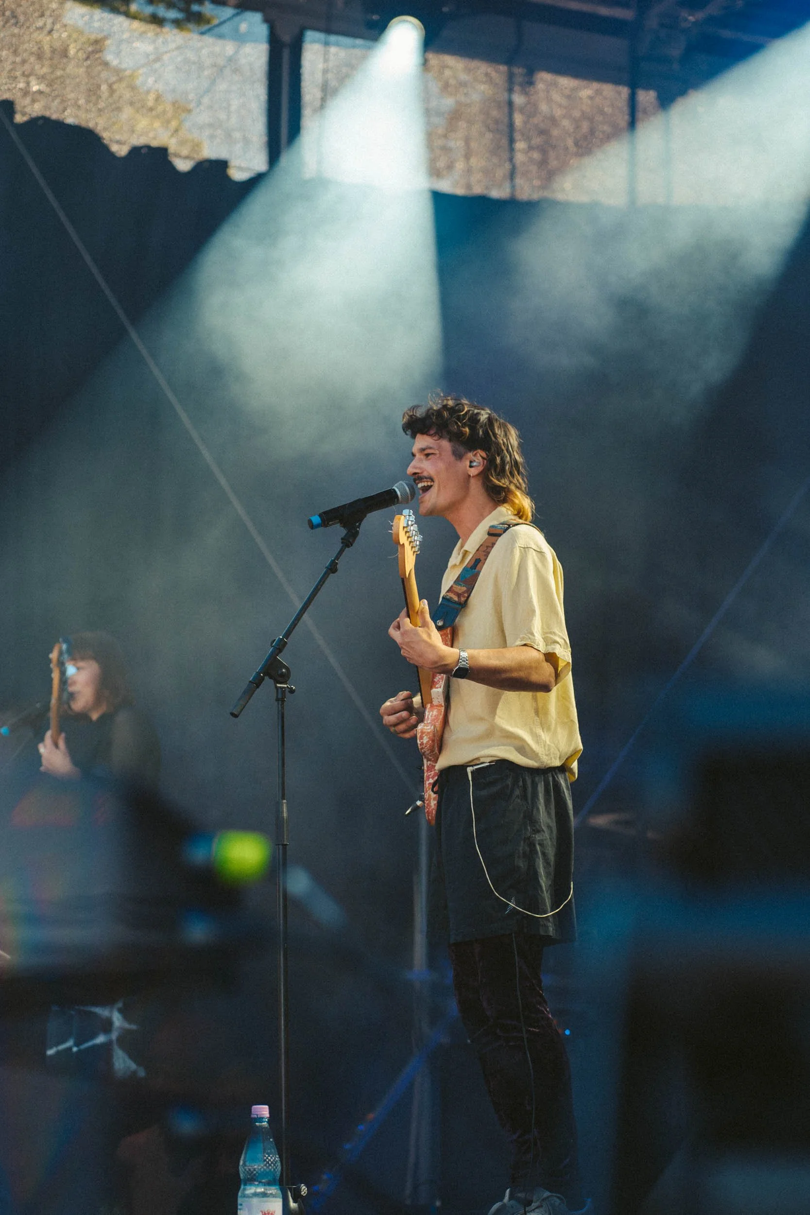 Ein junger Mann singt und spielt Gitarre auf einer Bühne mit Scheinwerfern, im Hintergrund eine weitere Musikerin, mit Wasserflasche auf dem Boden, während er lächelnd in ein Mikrofon singt.