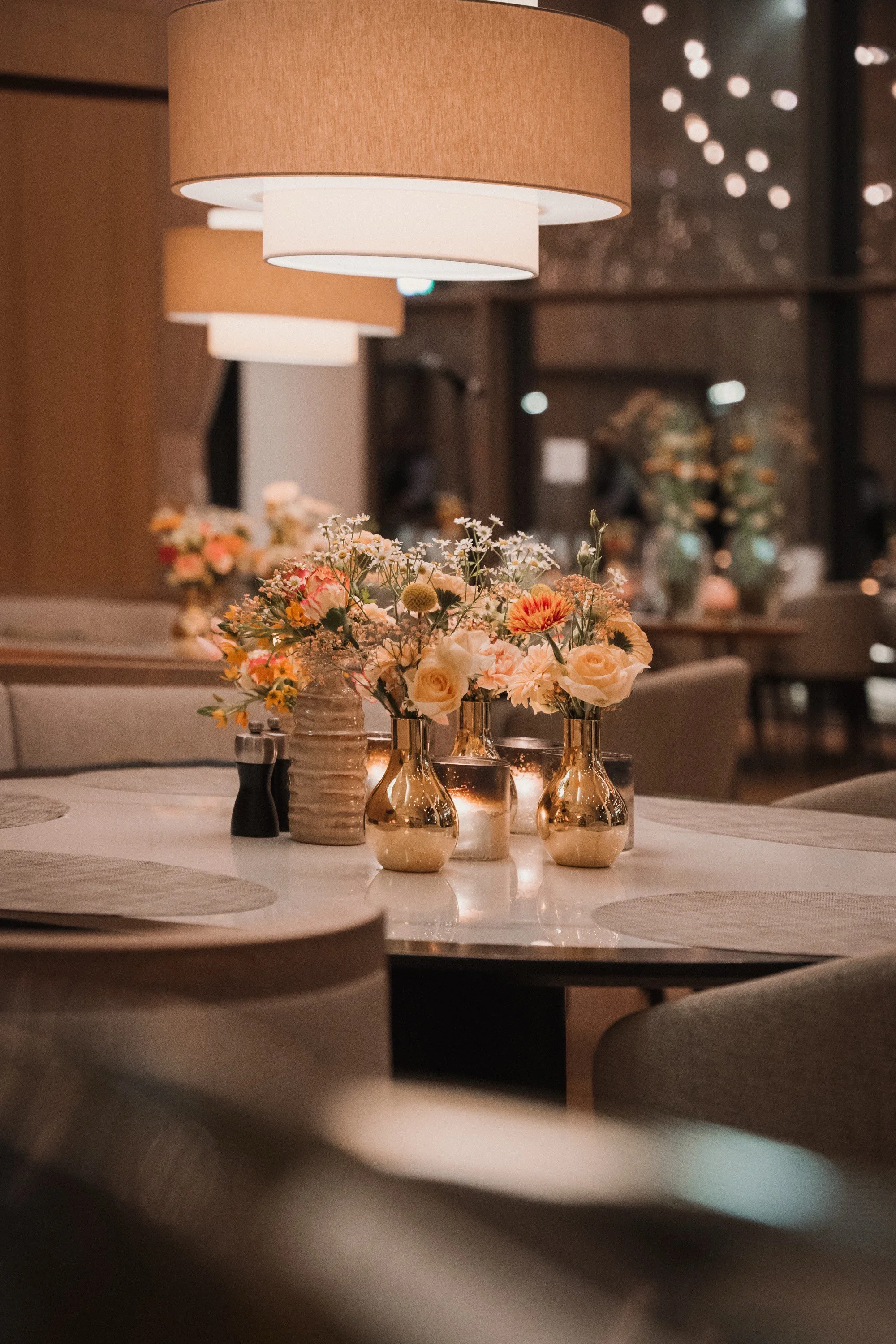 Ein gedeckter Esstisch mit Blumenarrangements in goldenen Vasen, einem Kerzenständer, Salz- und Pfefferstreuern, einem großen Lampenschirm und weiteren Blumen im Hintergrund in einem eleganten Restaurant.
