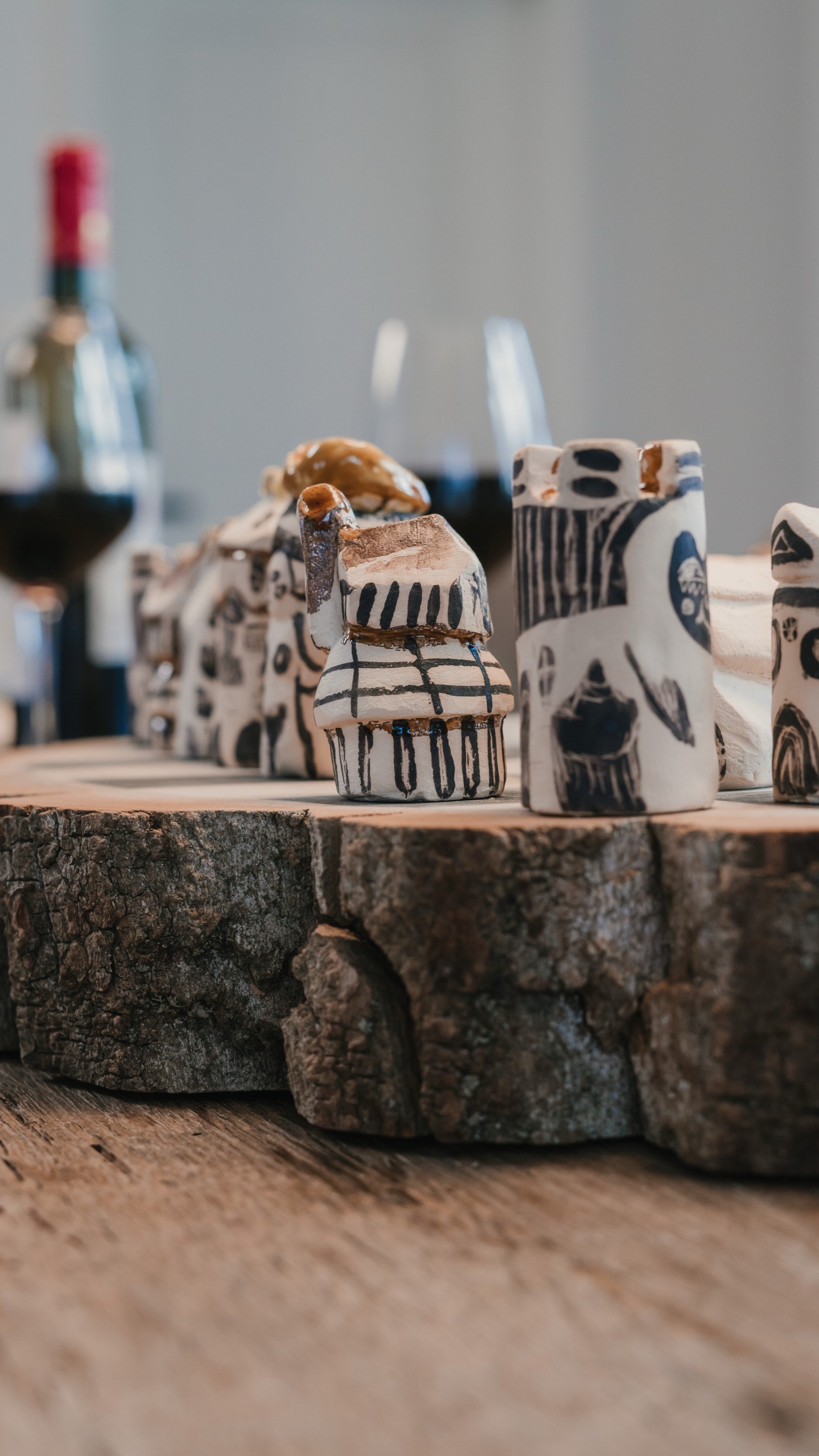 Kunstwerke aus Keramik mit schwarzen Linien und Mustern auf weißen Flächen, auf einem Holztisch. Getränke und Gläser im Hintergrund.