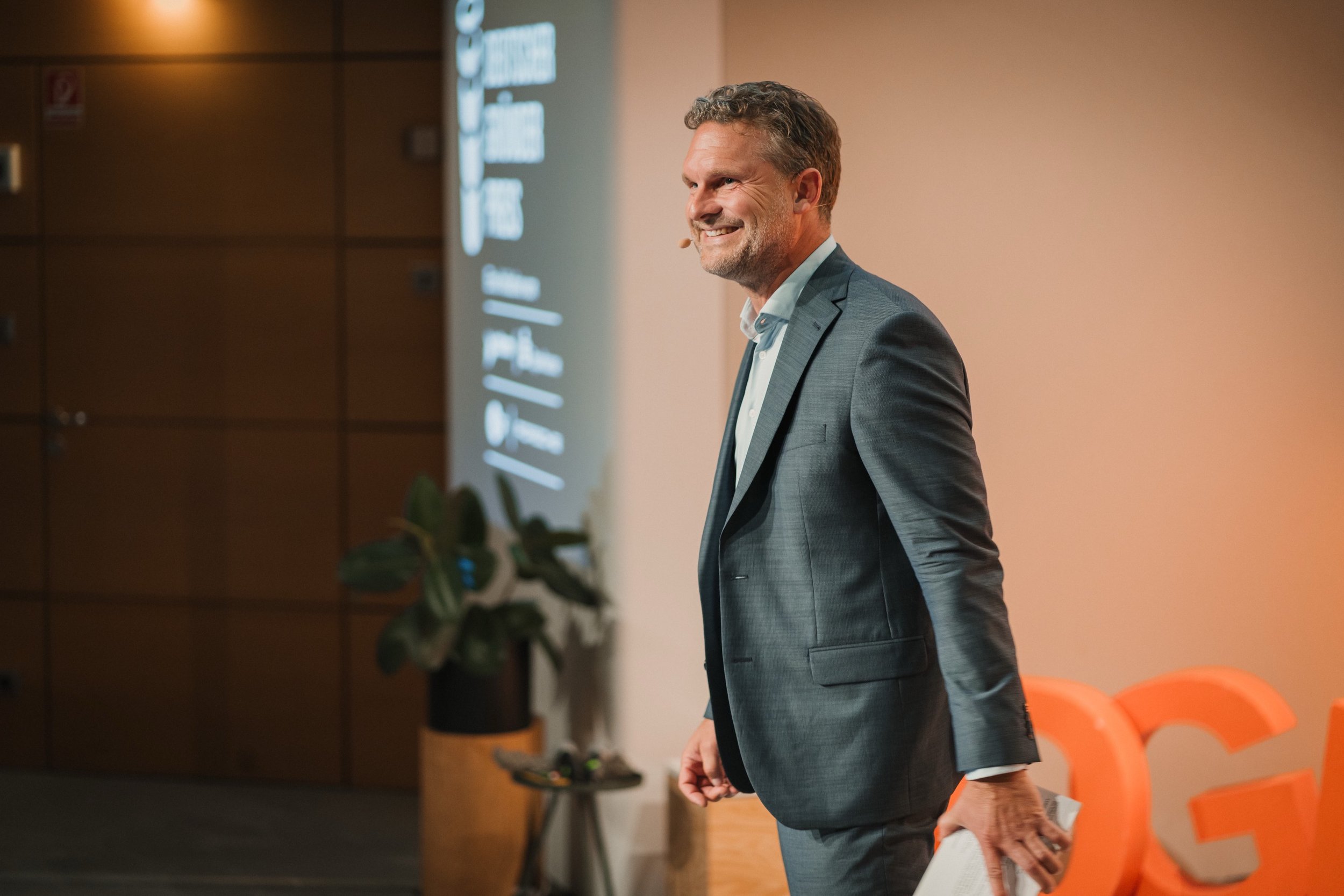 Ein Mann in einem grauen Anzug steht bei einer Präsentation, hält ein Blatt Papier in der Hand, lächelt und schaut nach links.