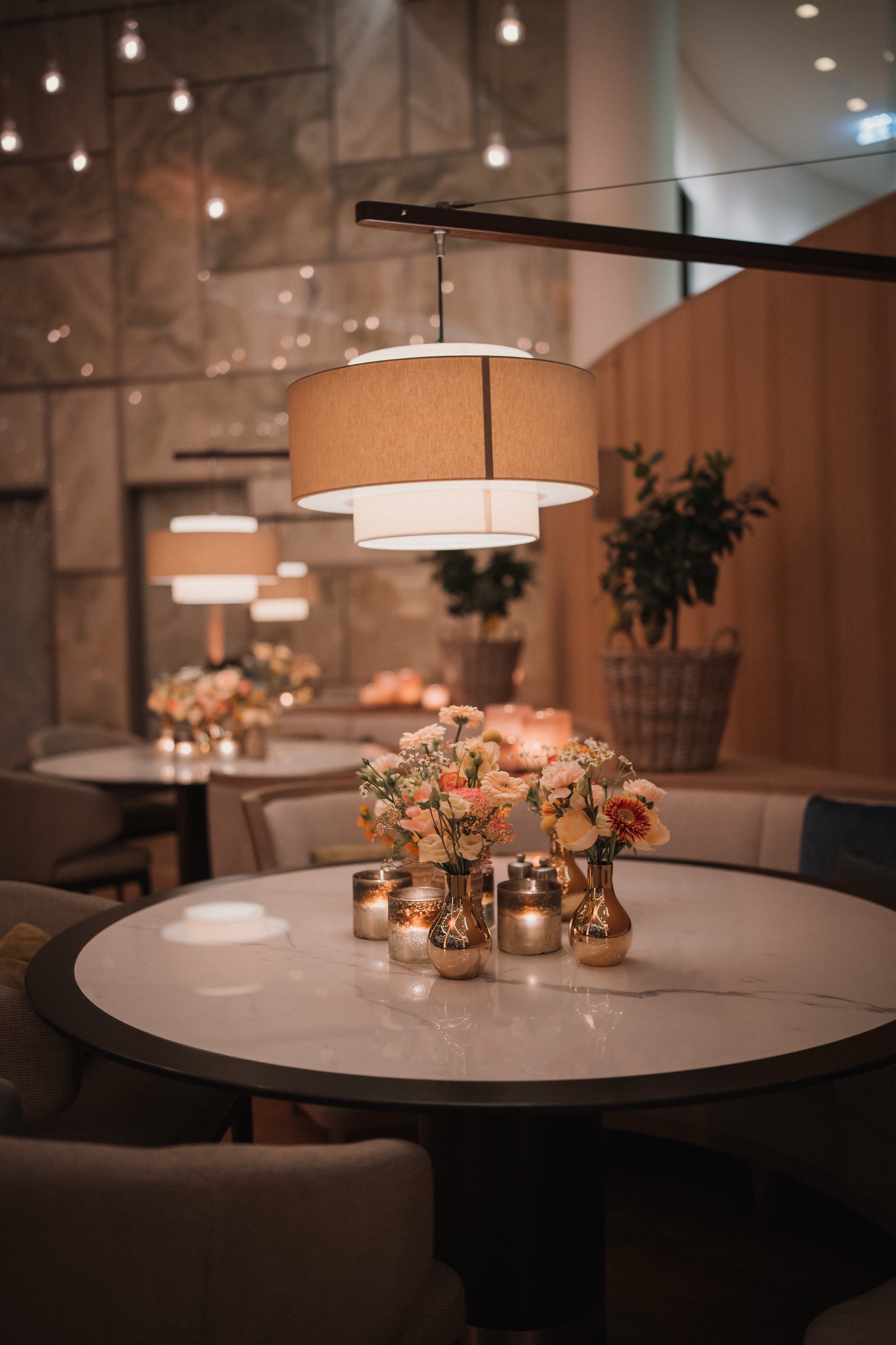 Gemütlich gedeckter Tisch mit Blumenvasen, Kerzen und einer großen Deckenlampe in einem eleganten Raum.