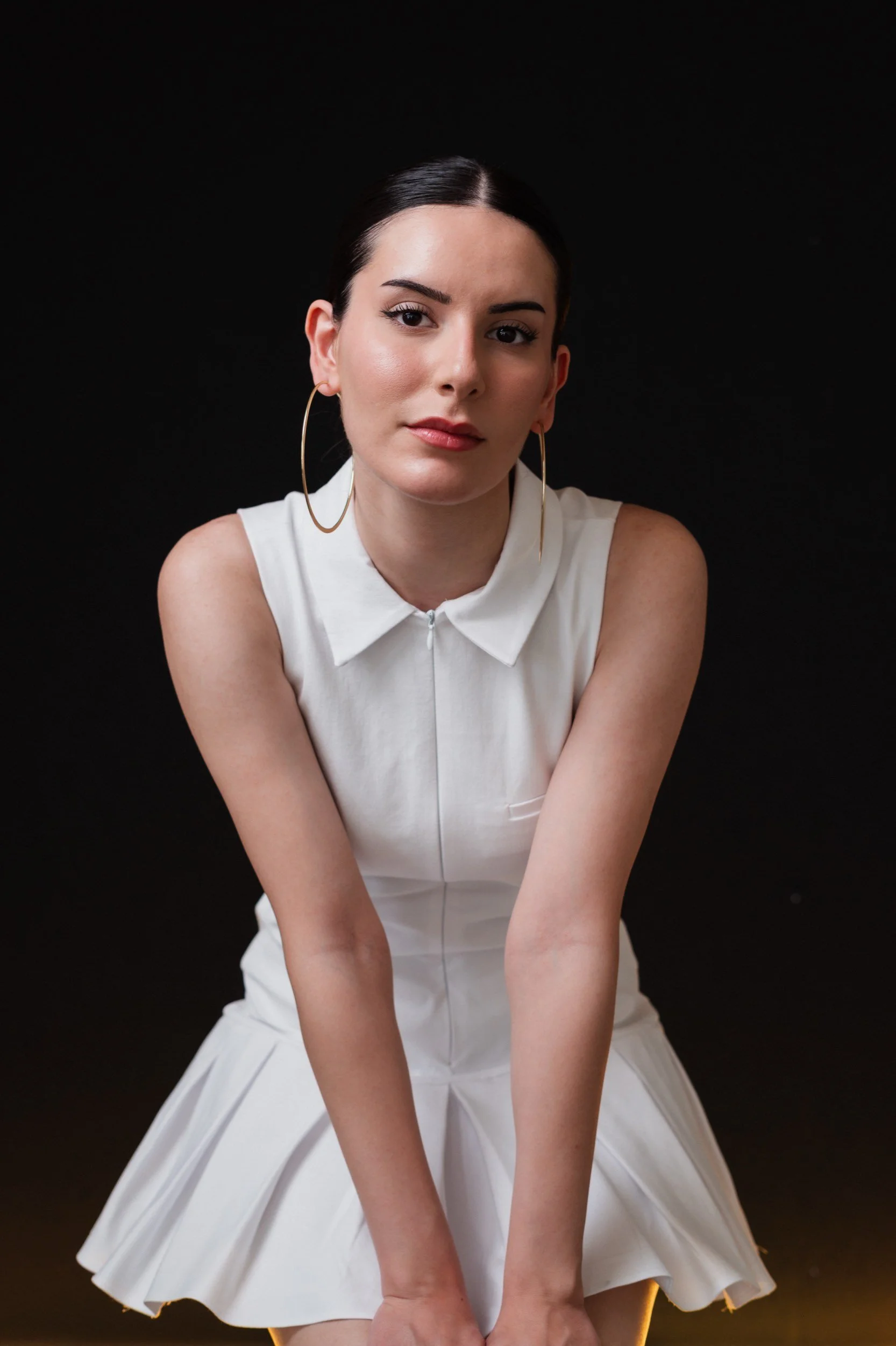Junge Frau mit dunklen Haaren, die eine weiße, ärmellose Bluse und einen weißen Rock trägt, sitzt vor schwarzem Hintergrund.