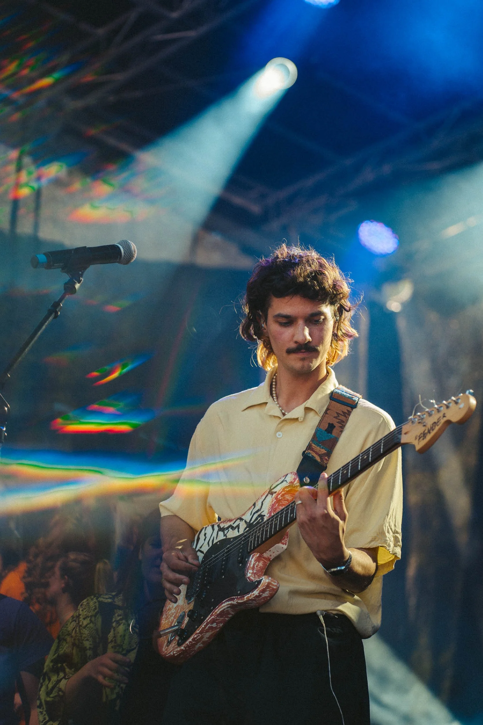 Ein junger Mann spielt auf einer E-Gitarre während eines Konzerts, mit bunten Lichtern und einer Bühne im Hintergrund.
