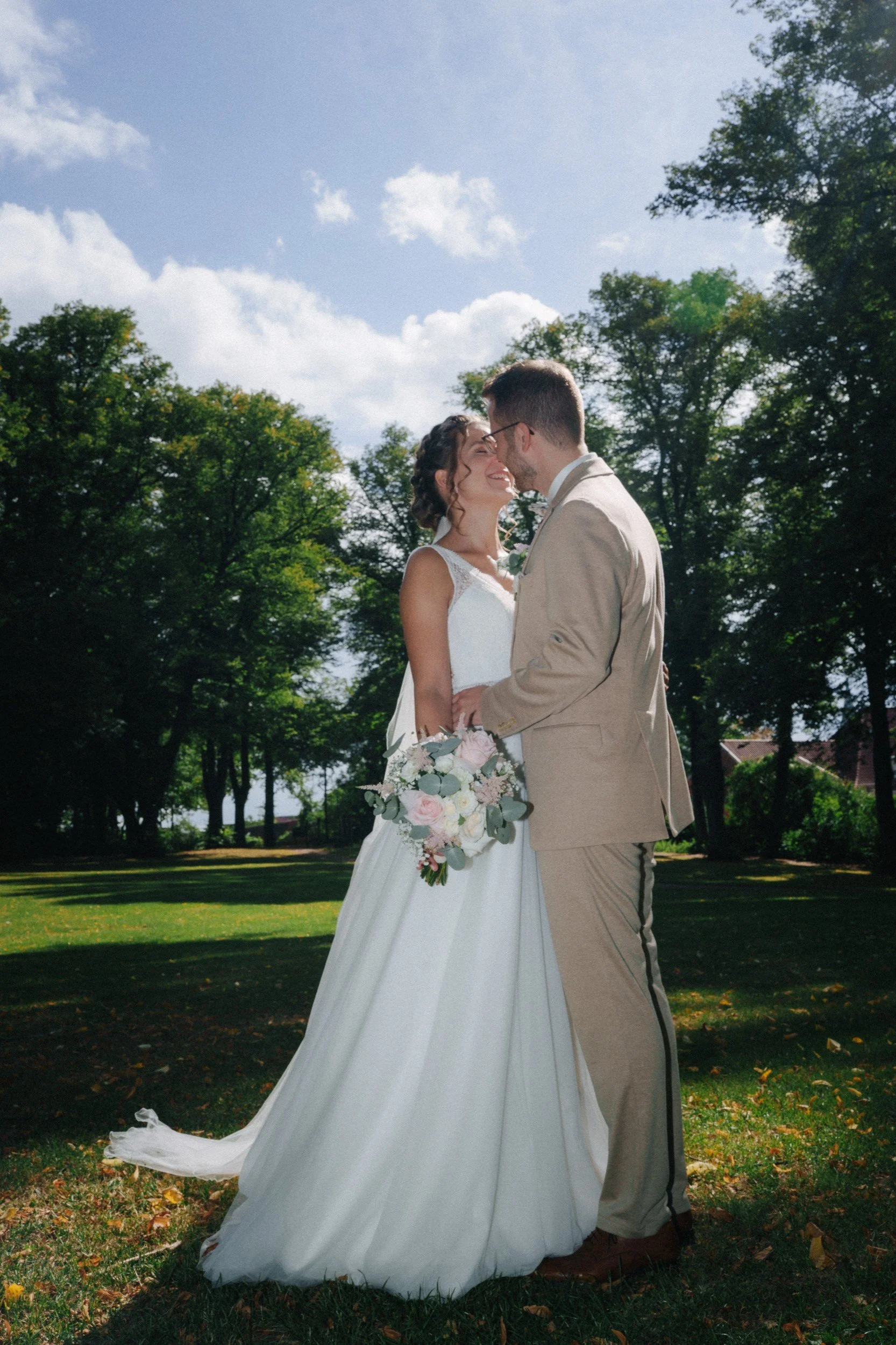 Ein Brautpaar umarmt sich im Freien an einem sonnigen Tag, im Hintergrund Bäume und blauer Himmel.
