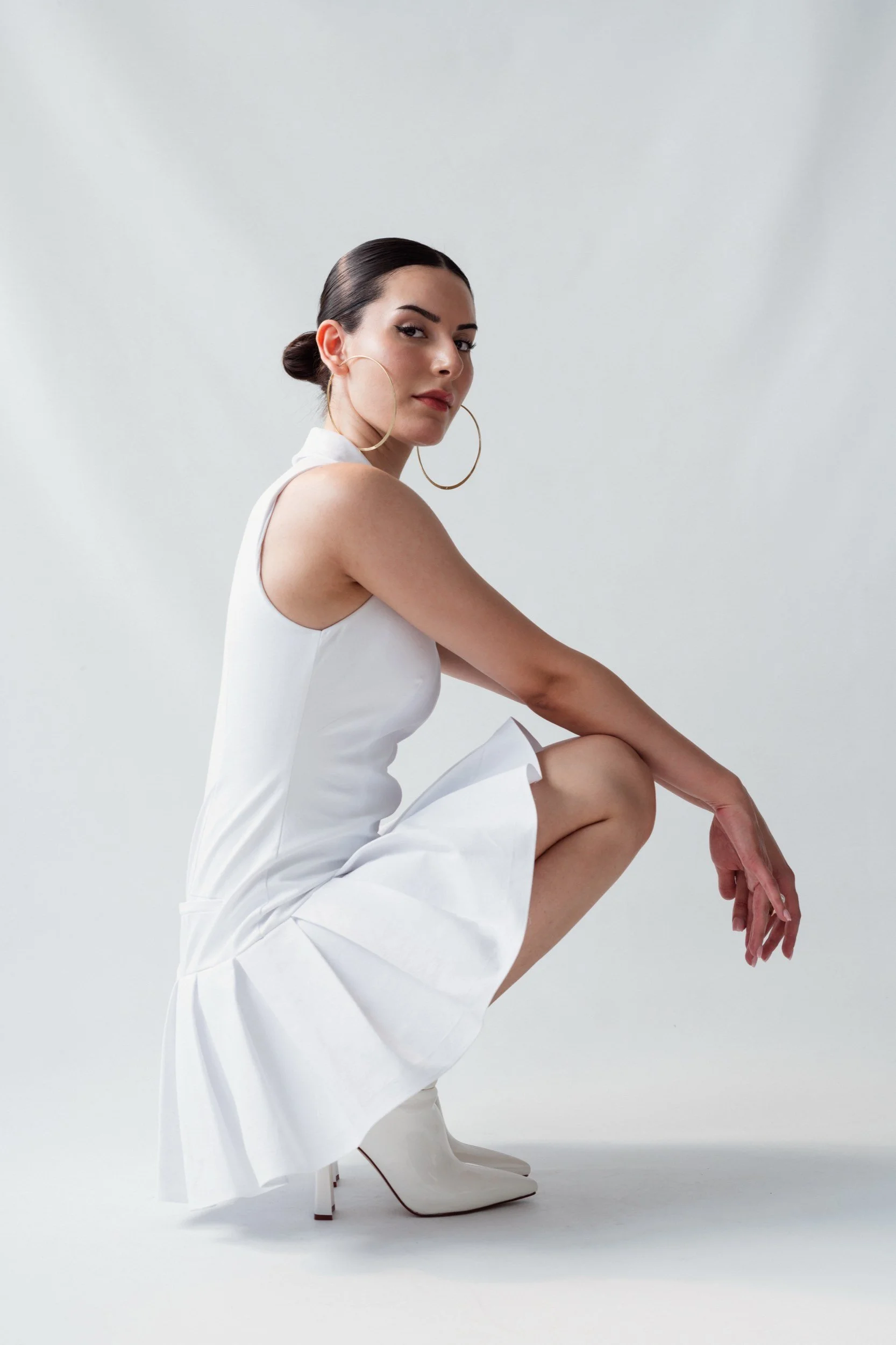 Eine Frau in weißem Kleid und weißen Stiefeln mit großen goldenen Ohrringen sitzt in die Hocke auf einer weißen Studio-Hintergrund.