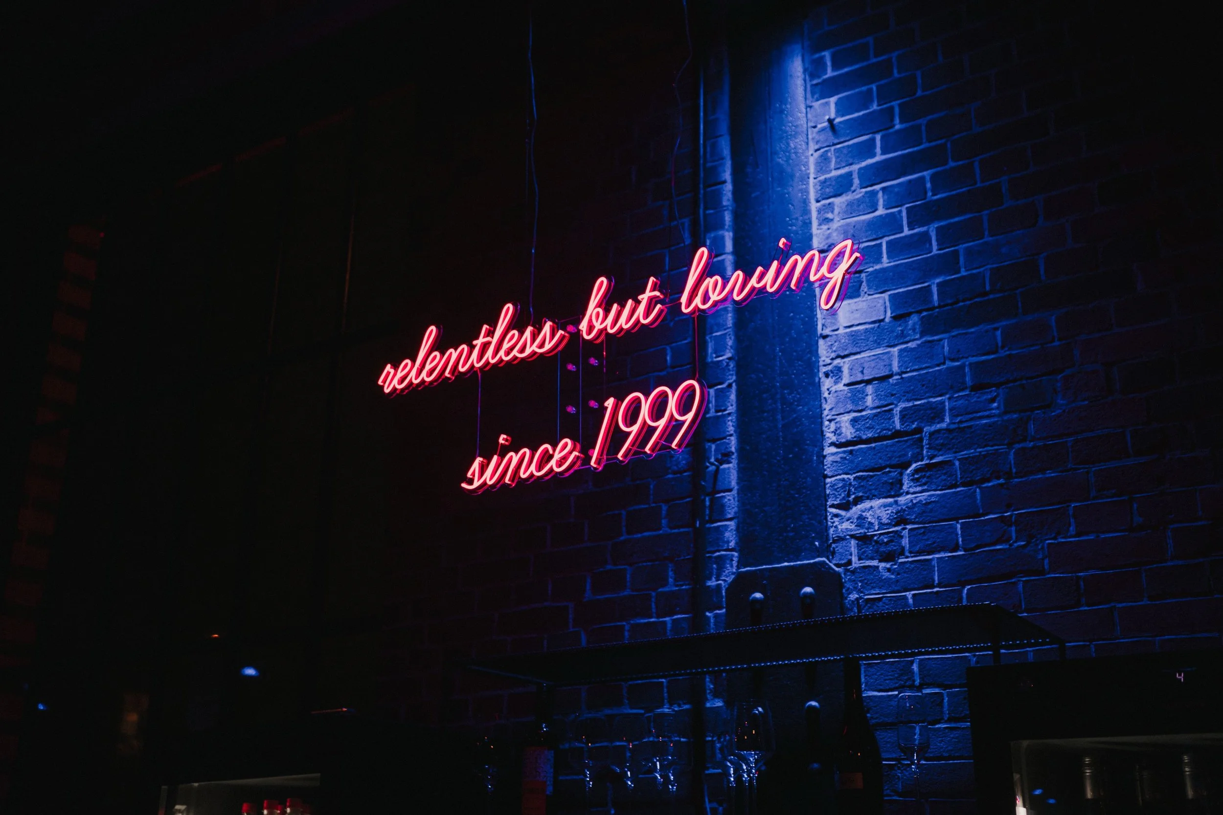 Neon-Leuchtreklame an einer dunklen Ziegelmauer mit der Aufschrift "relentless, but loving since 1999" in Pink.