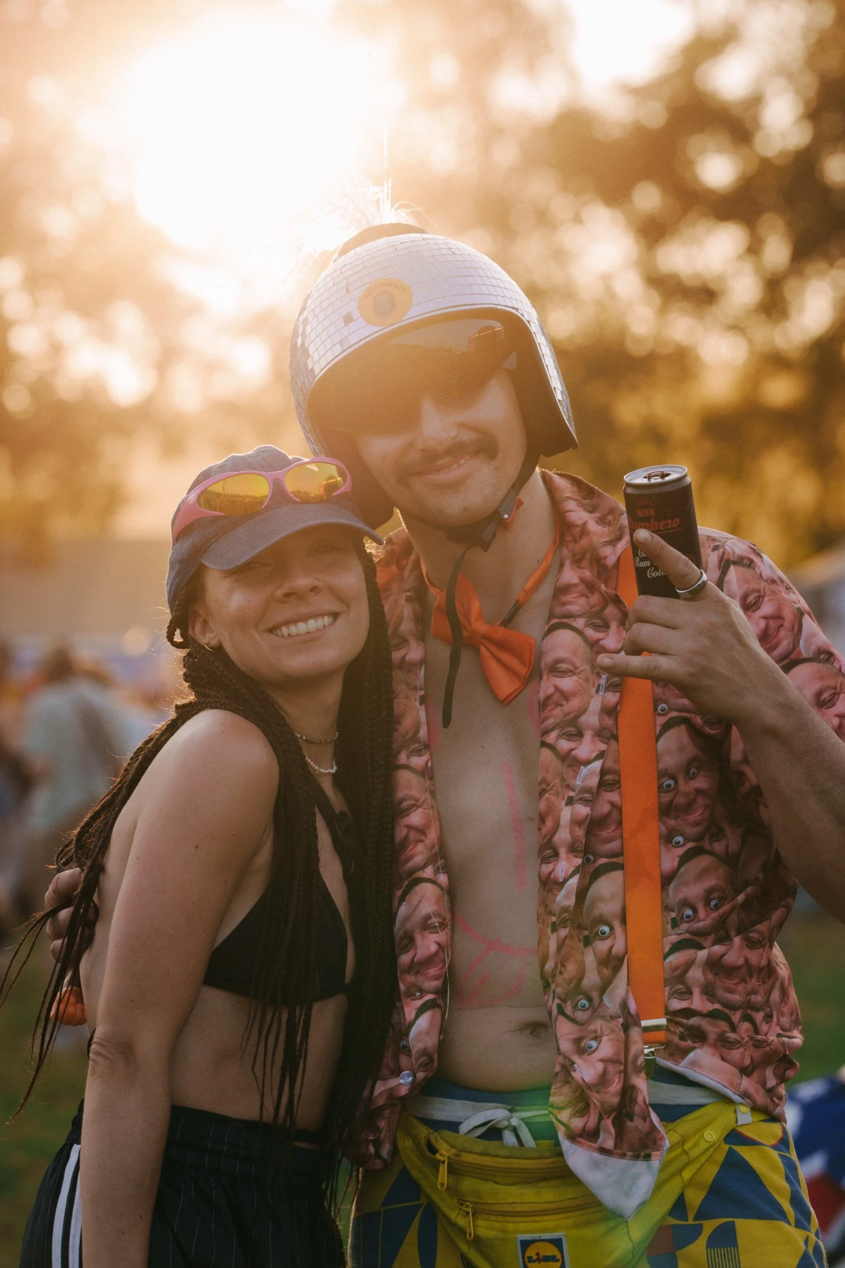 Zwei Menschen bei einem Outdoor-Event, Sonnenlicht im Hintergrund, feiern, lächeln, tragen sommerliche Kleidung und Accessoires wie Sonnenbrillen, ein Mann trägt einen Helm.