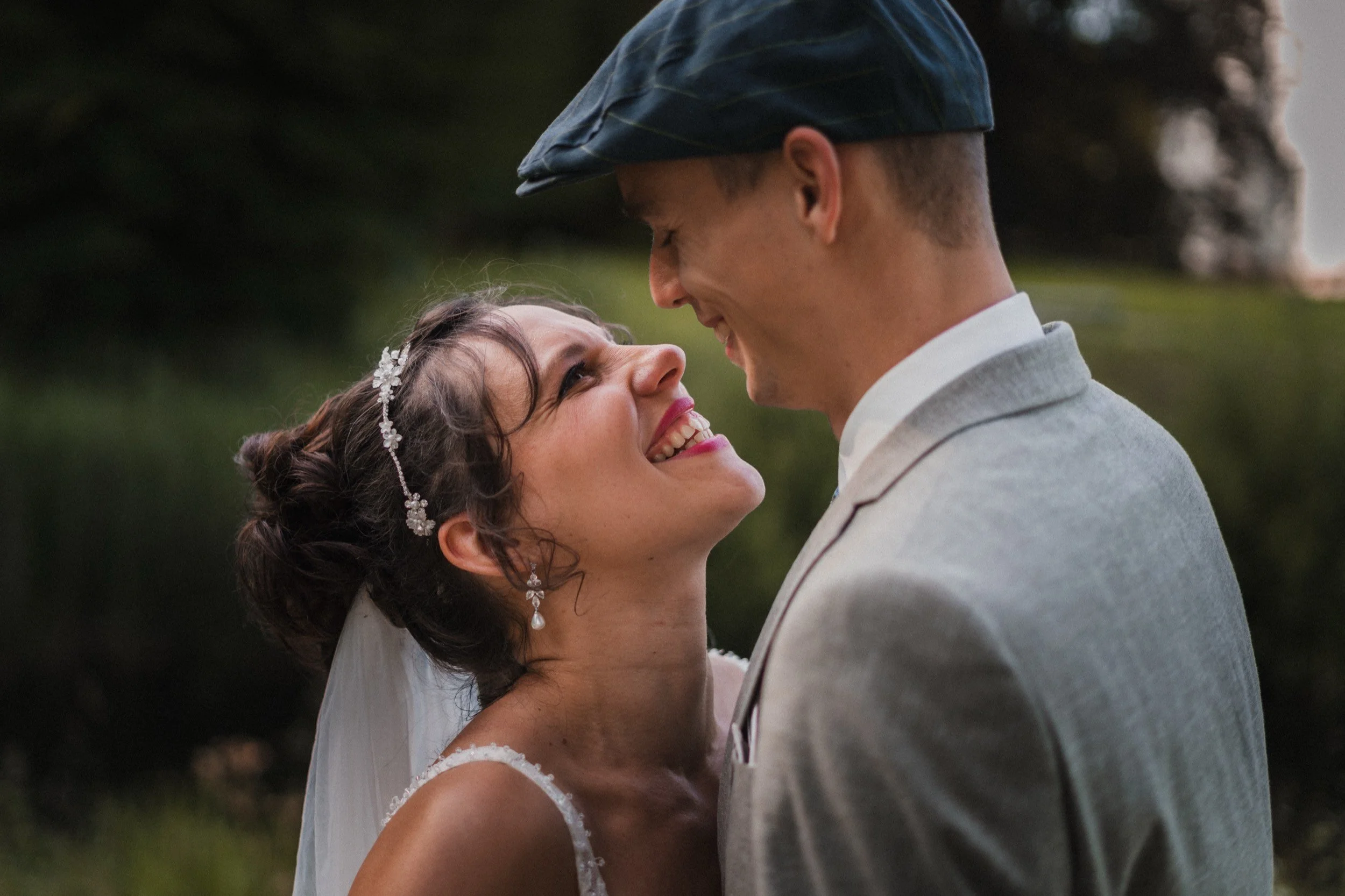 Ein Brautpaar beim Blick in die Augen vor einem Naturhintergrund, die Braut trägt einen weißen Schleier und Schmuck, der Bräutigam trägt einen hellgrauen Anzug.