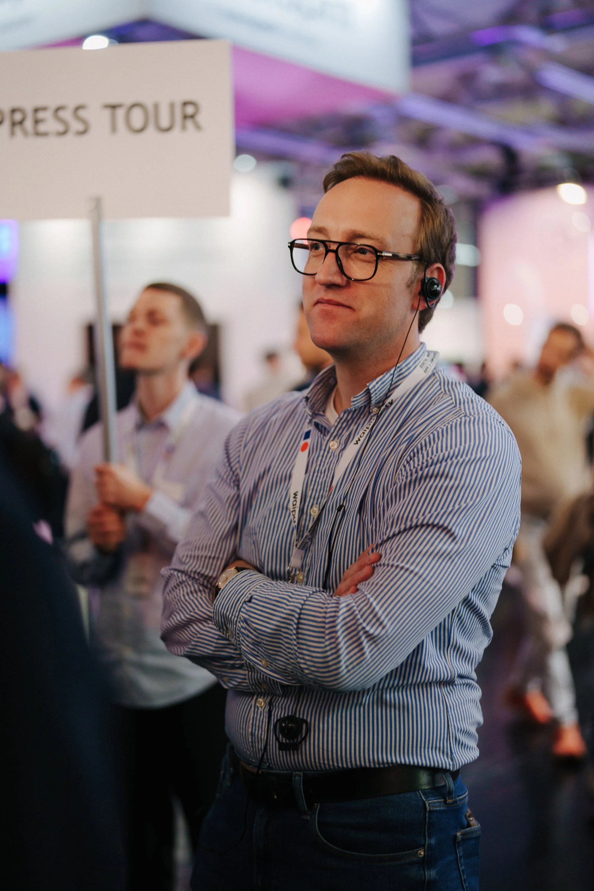 Ein Mann mit Brille, gepunktetem Hemd und Kopfhörer steht mit verschränkten Armen bei einem Messe- oder Konferenz-Event.