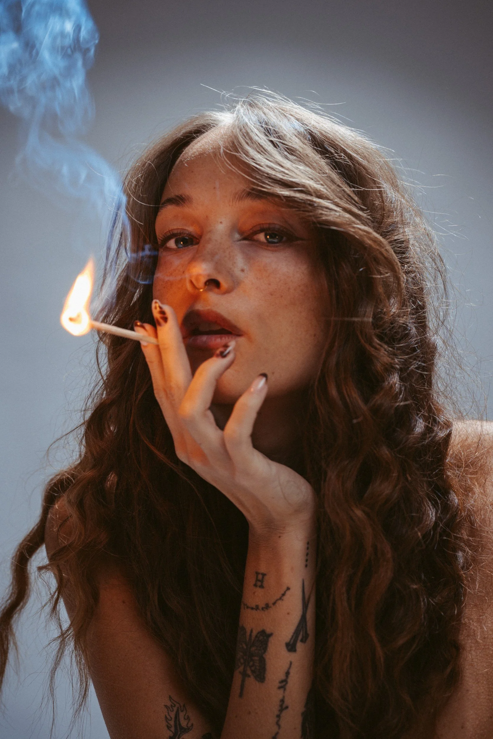 Junge Frau mit Tattoos im Arm hält eine brennende Zigarette in der Hand, Rauch steigt auf, Blick direkt in die Kamera, grauer Hintergrund.