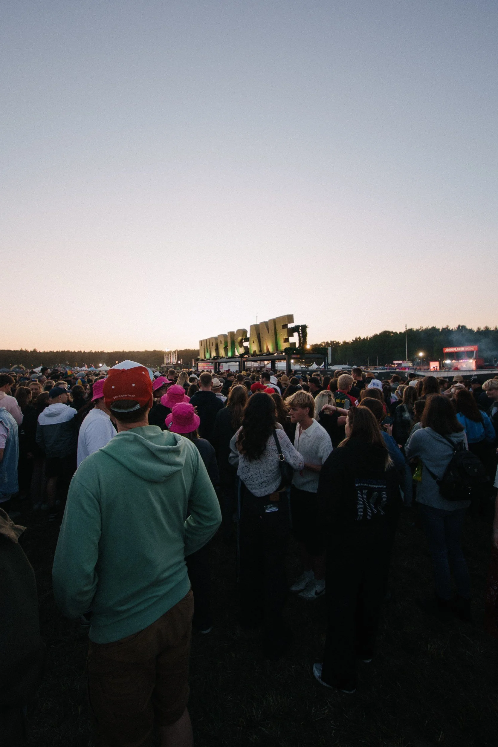 Menschenmenge bei einem Konzert im Freien mit einem großen Schild, das 'HURRICANE' sagt, bei Sonnenuntergang.
