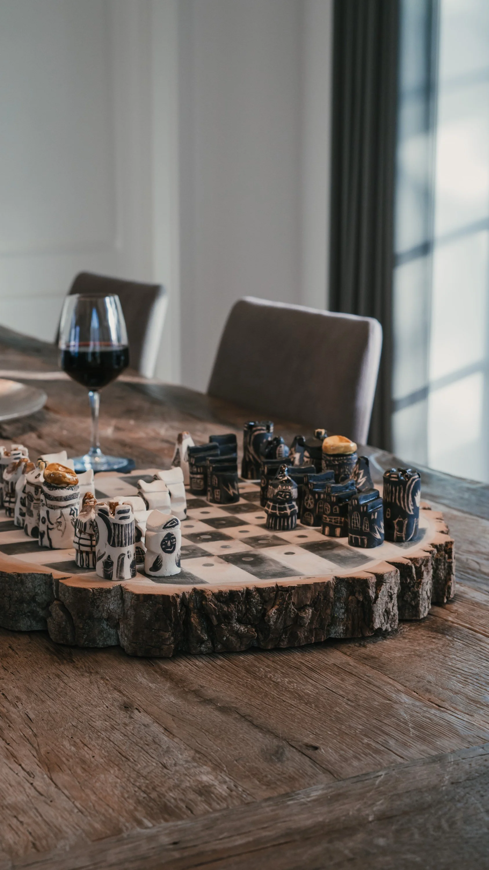 Schachspiel auf einem runden Holzbrett mit schwarzer und weißer Figuren auf einem Holztisch, im Hintergrund ein Glas Rotwein und ein Fenster mit dunklen Vorhängen.