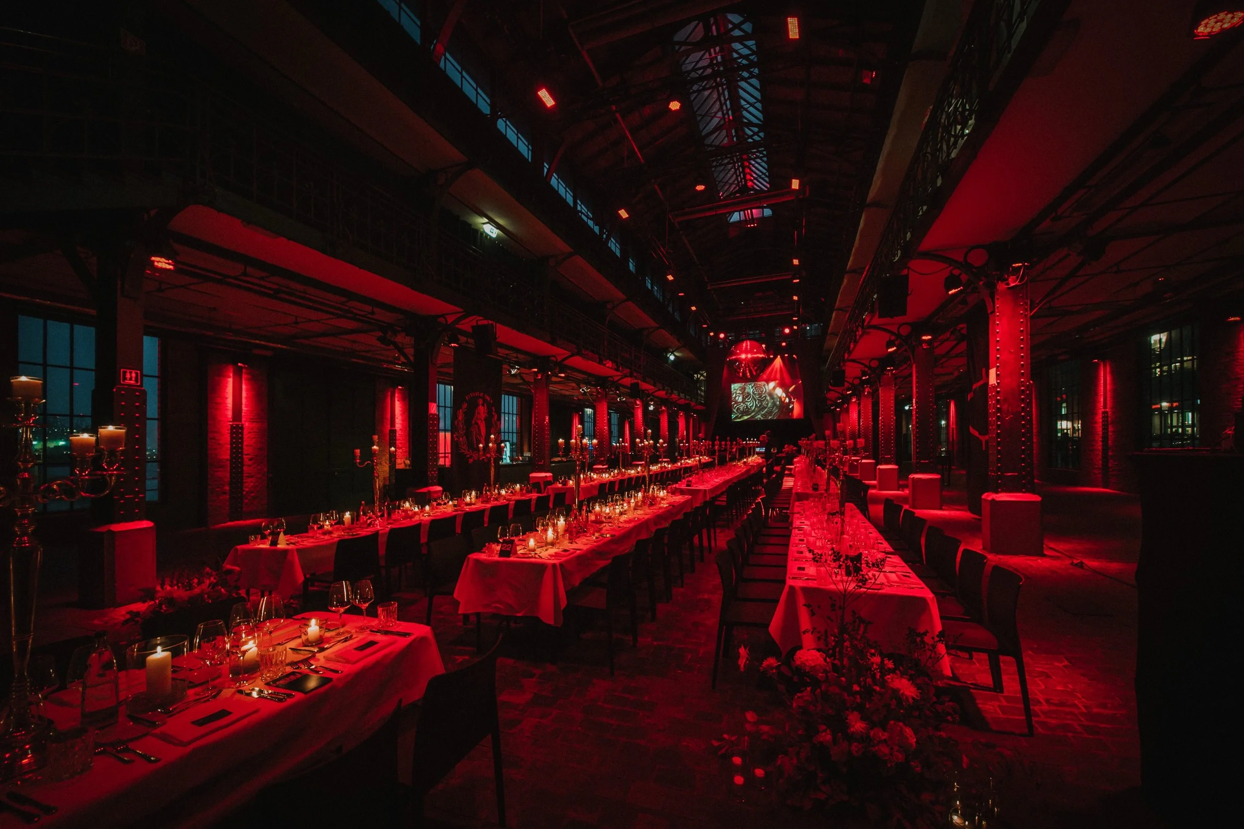 Ein eleganter Festsaal mit langen, gedeckten Tischen, Kerzenlicht, roter Beleuchtung, Blumen und dekorativen Elementen, bereit für eine Veranstaltung.