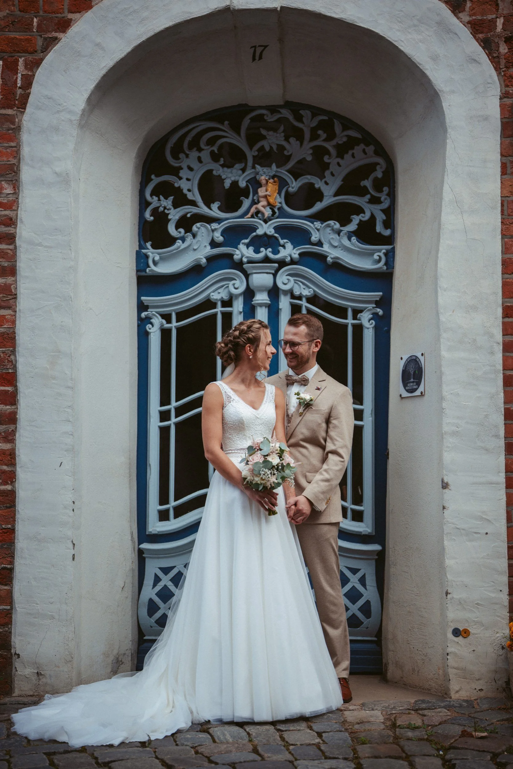 Ein Brautpaar steht vor einer kunstvollen blauen Tür, die in eine weiße Mauer eingelassen ist. Die Braut trägt ein weißes Hochzeitskleid und hält einen Blumenstrauß, während der Bräutigam in einem hellbraunen Anzug mit Fliege lacht. Das Paar blickt s