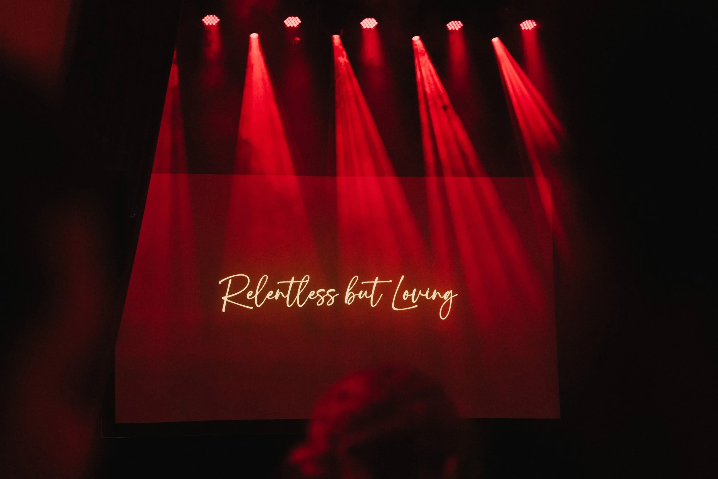 Auf einer LED-Leinwand ist der Text 'Relentless but Loving' zu sehen, beleuchtet durch rote Scheinwerfer in dunkler Umgebung.