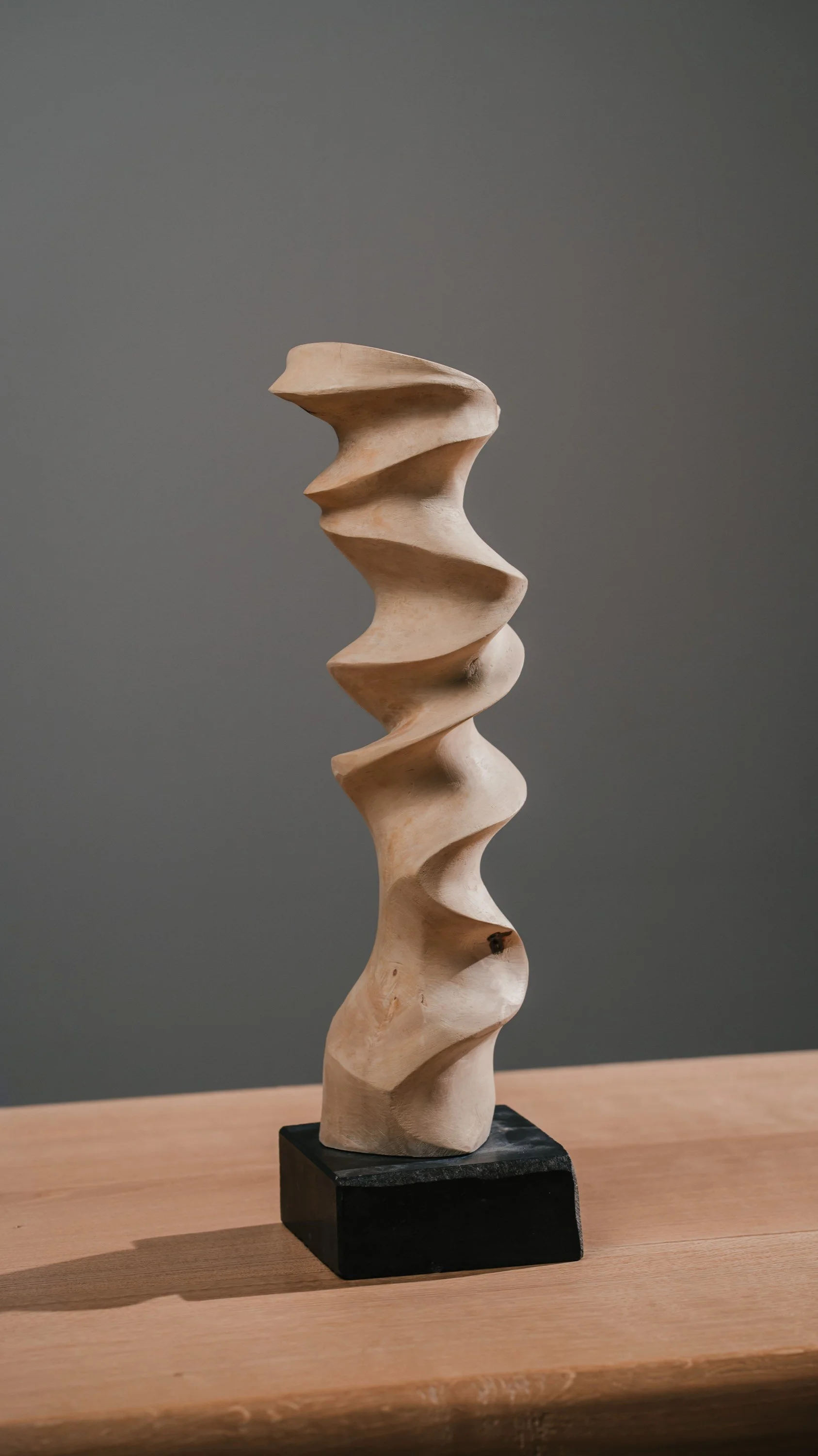 Studio-Fotografie von einem Kunstwerk. Skulptur aus Holz auf schwarzem Sockel, mit spiralförmigen, geschwungenen Formen