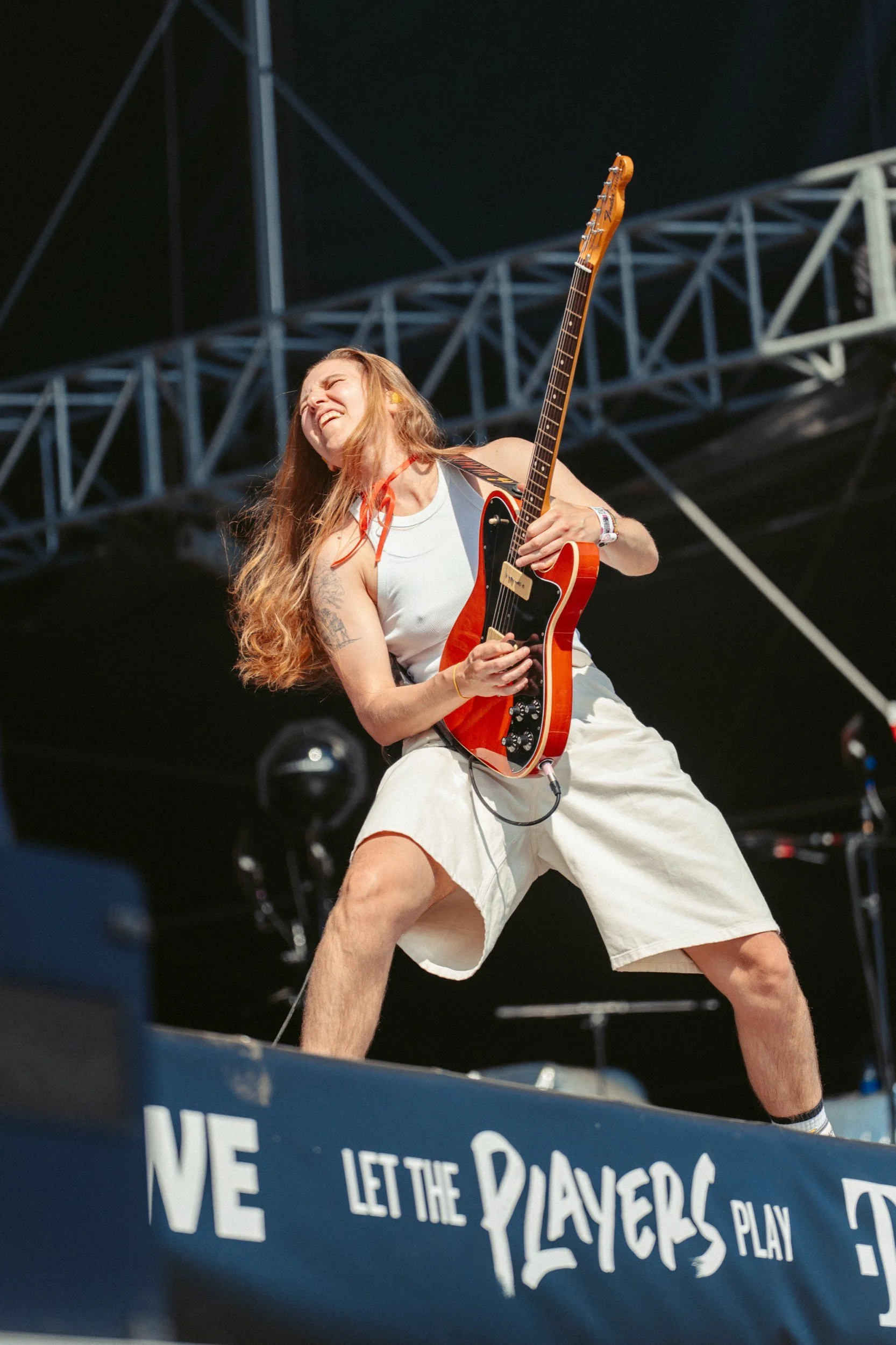 Junge Frau spielt elektrische Gitarre auf einer Bühne während eines Konzerts, trägt weiße Kleidung, hat lange Haare und zeigt eine leidenschaftliche Pose.