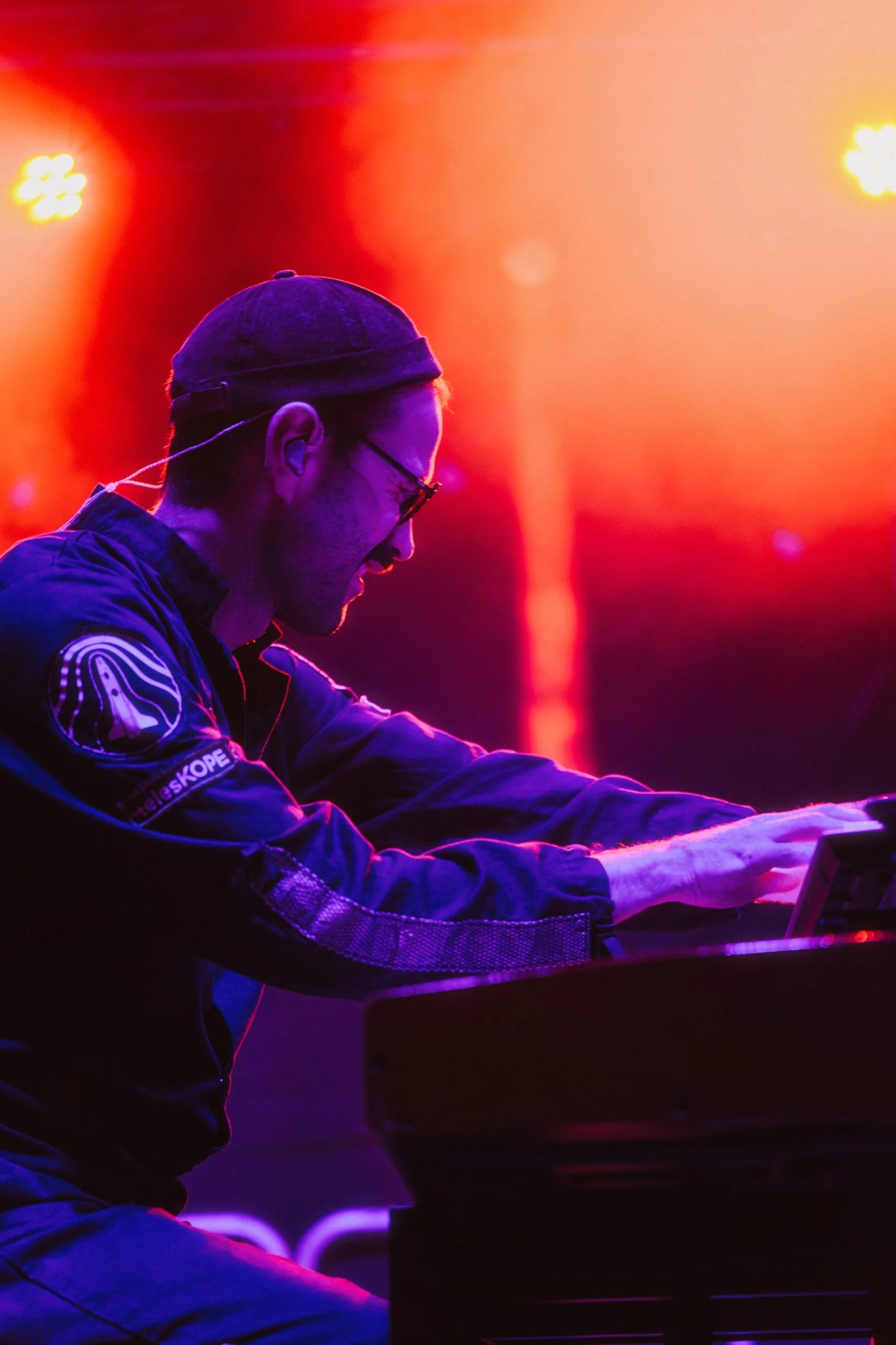 Ein Mann spielt ein Keyboard auf einer Bühne, beleuchtet von bunten LED-Lichtern, trägt eine schwarze Jacke, eine schwarze Mütze und eine Brille.