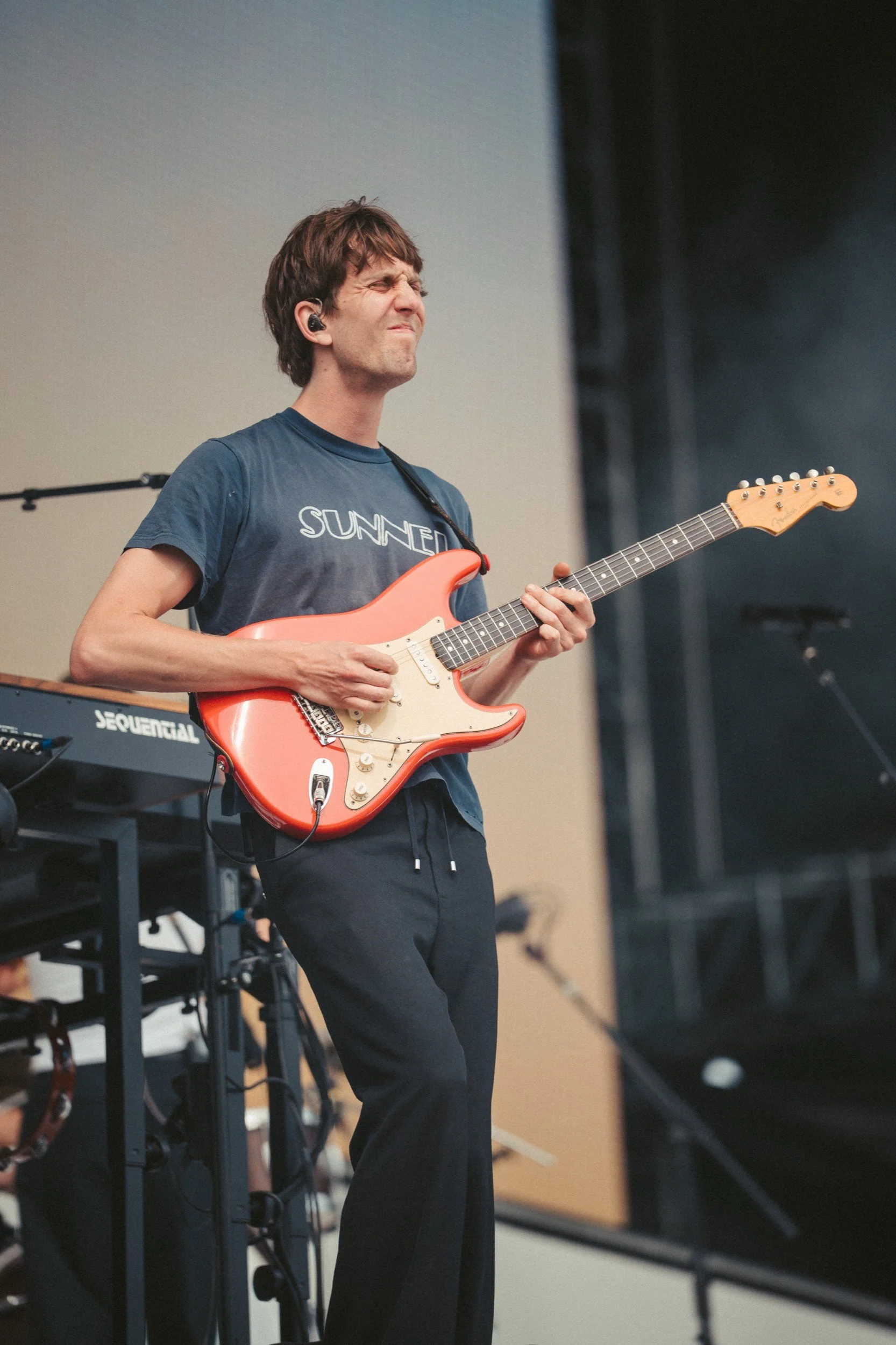 Ein junger Mann spielt auf einer Bühne E-Gitarre. Er trägt ein T-Shirt mit dem Schriftzug "Sunset" und scheint beim Spielen emotional zu sein.
