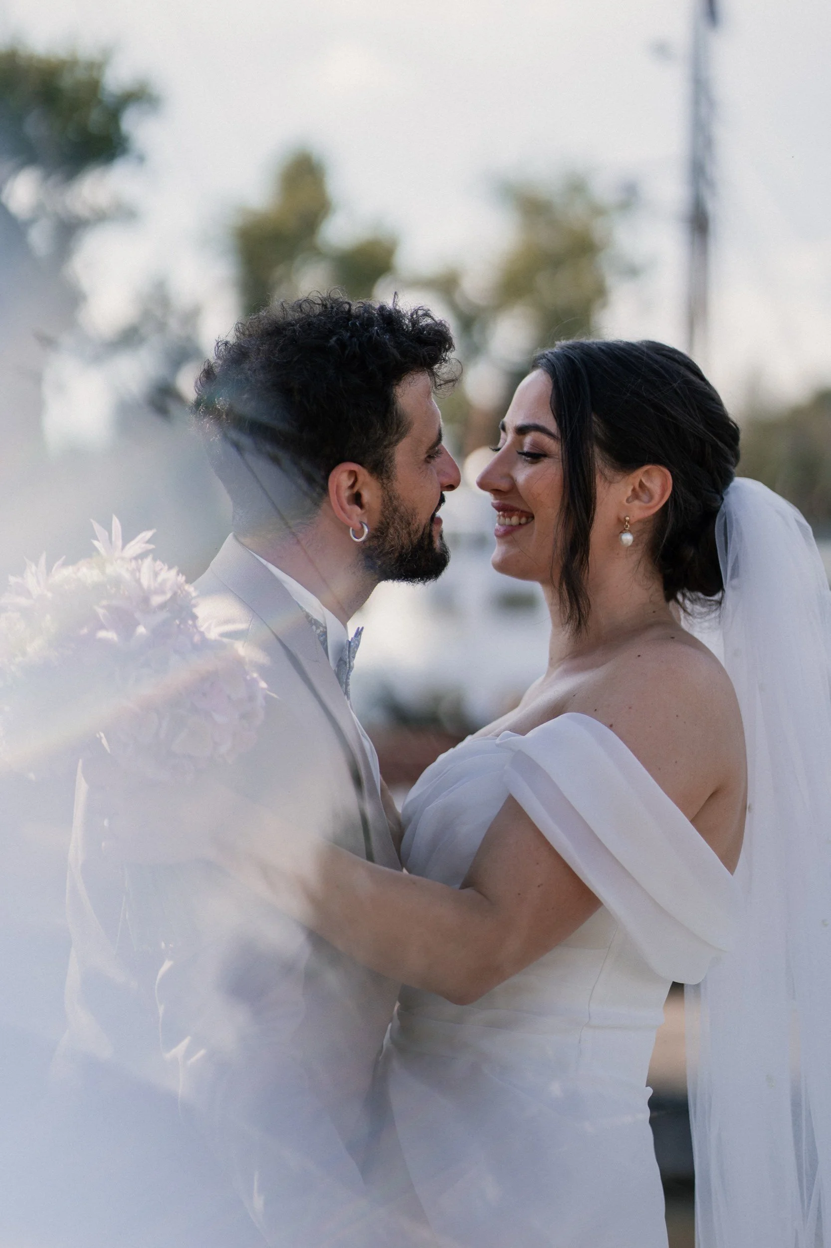 Ein Brautpaar steht sich im Freien gegenüber, lächelt und ist emotional verbunden bei einer Hochzeit, mit Bäumen und einem Himmel im Hintergrund.