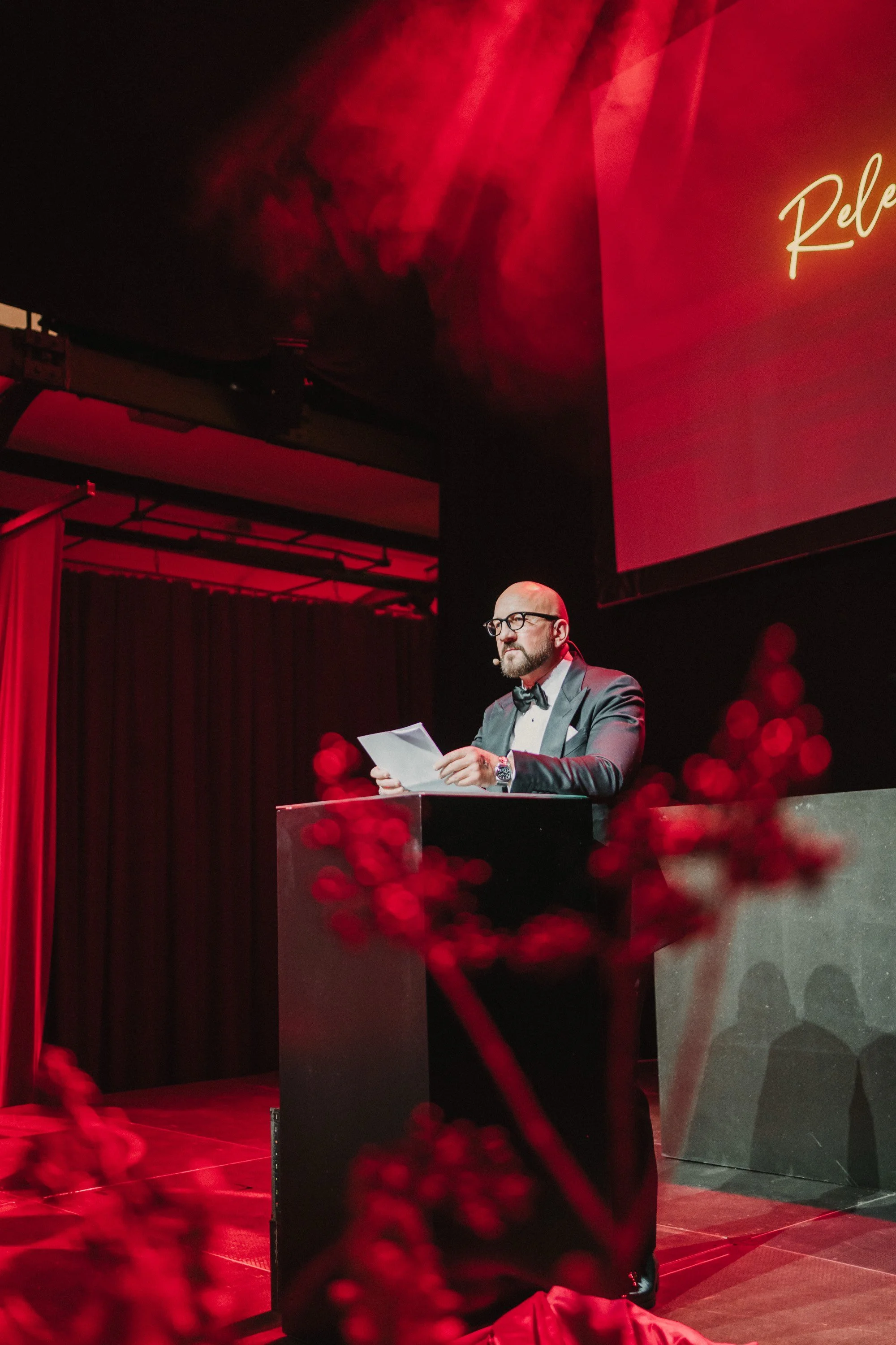 Ein Mann in formeller Kleidung steht an einem Podium bei einer Veranstaltung, liest aus Papieren, mit rotem Beleuchtung und einem großen Bildschirm im Hintergrund.