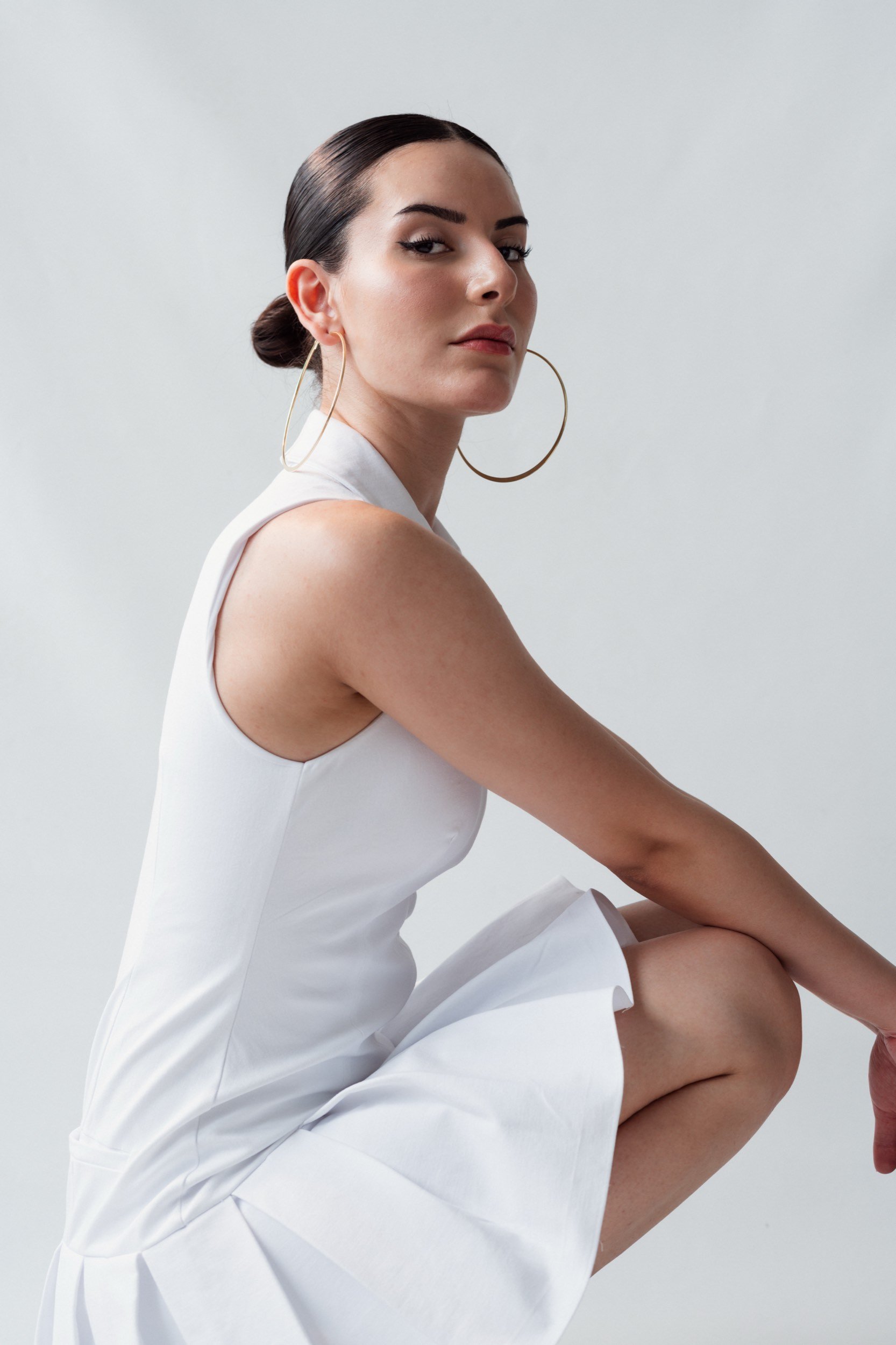 Junge Frau in Weißes Kleid mit großen Ohrringen in einer sitzenden Pose auf einer leichten grauen Hintergrund.