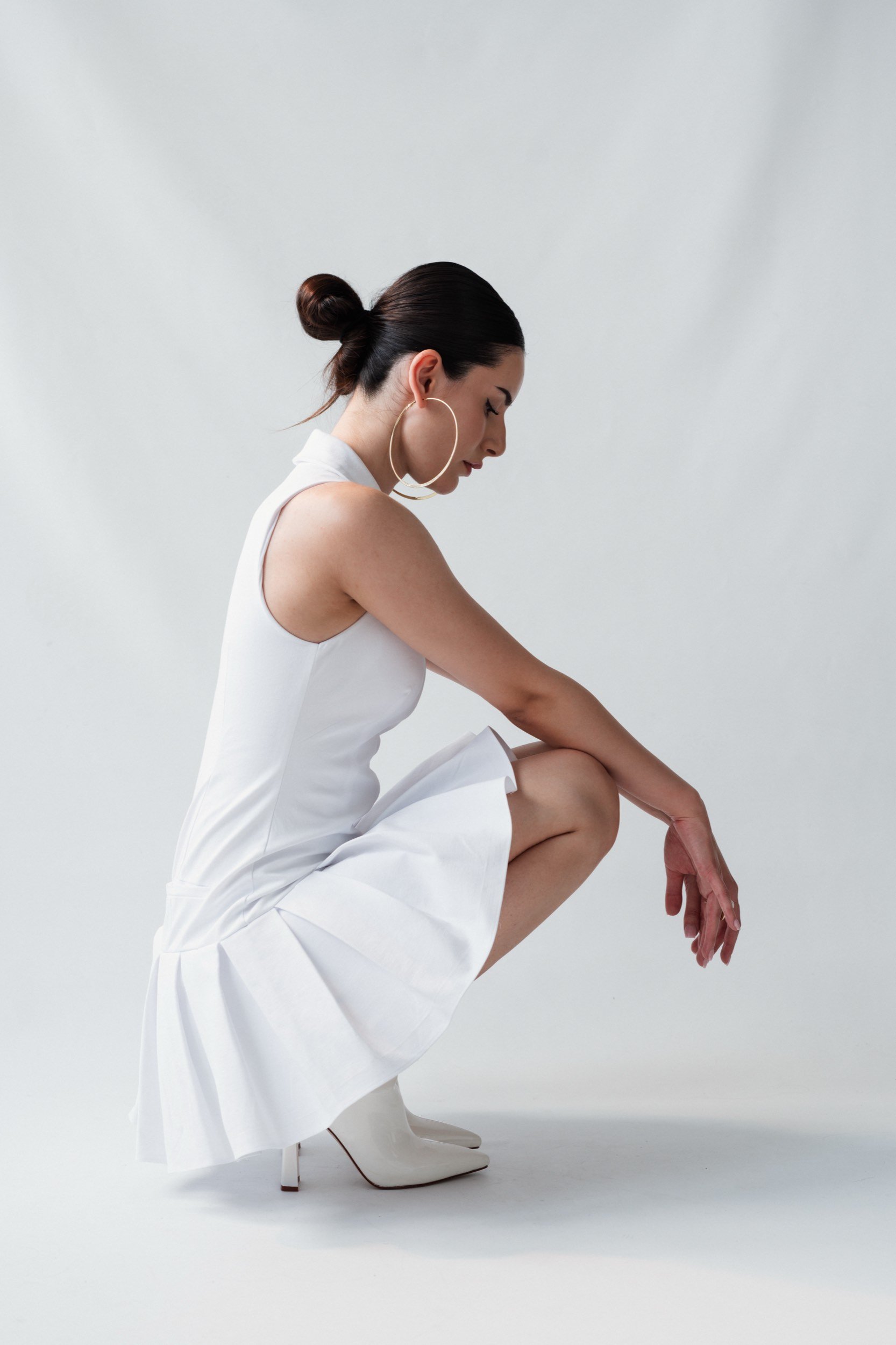 Eine Frau in einem weißen Kleid und hohen weißen Schuhen ist in die Hocke gegangen, mit einer Hand an ihrem Knie und der anderen Hand nach unten. Sie trägt große Ohrhörer und hat dunkles, glattgezogenes Haar zu einem Knoten gebunden.