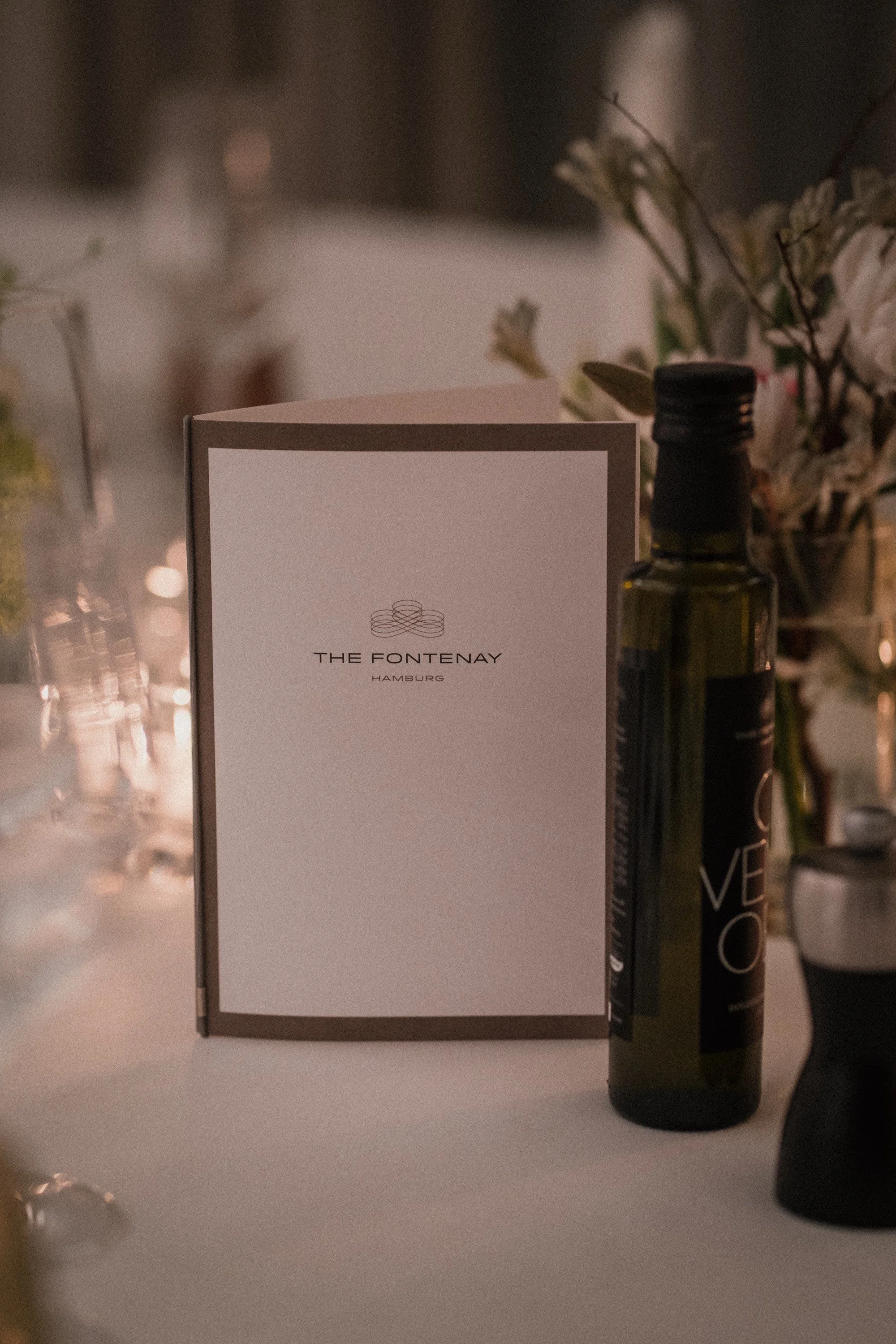 Menükarte von THE FONTENAY Hamburg auf einem gedeckten Tisch, umgeben von Blumen und einem Flaschenöl.