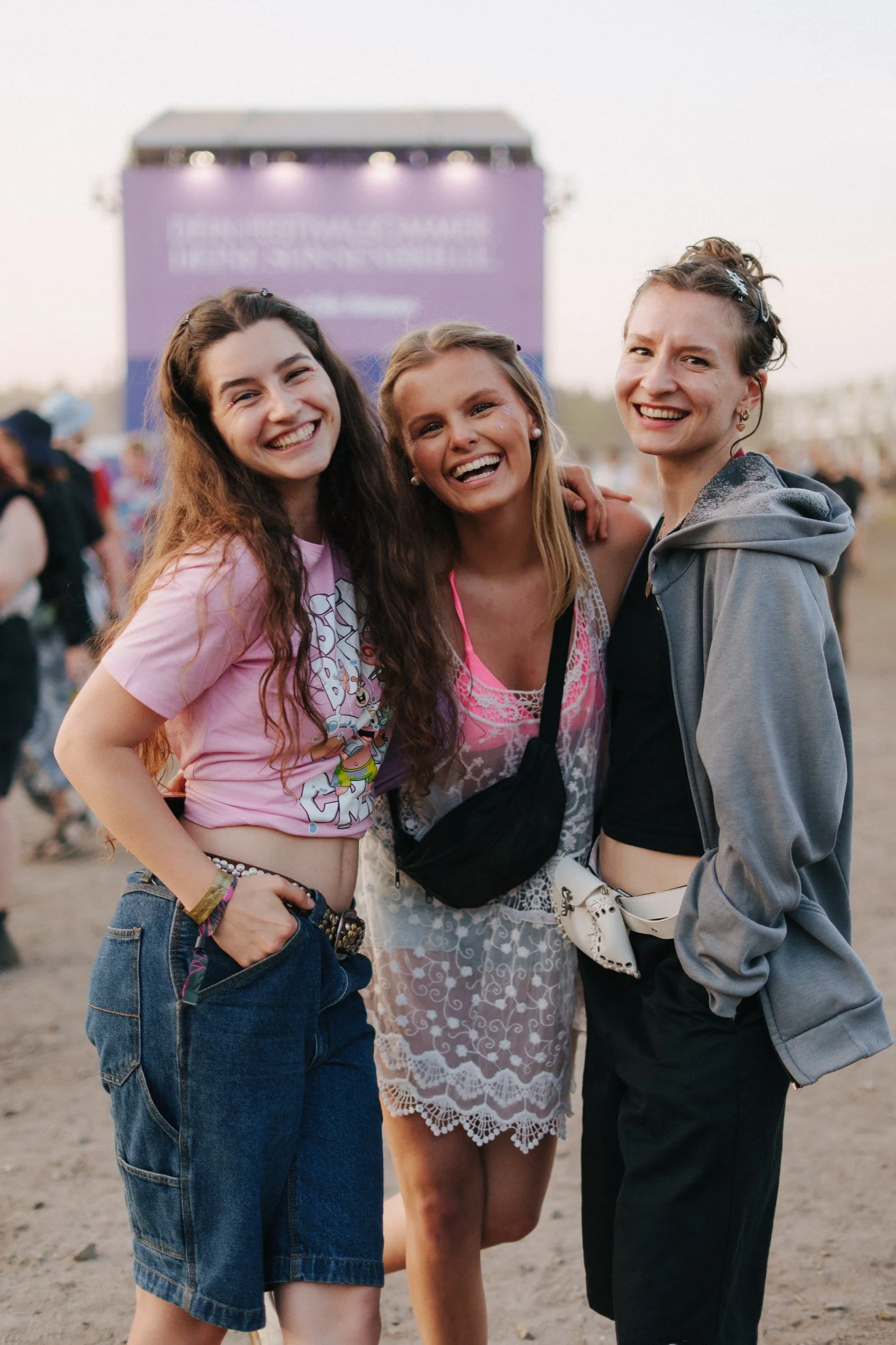 Drei lachende junge Frauen beim Festival im Freien, umgeben von anderen Menschen und einer großen Bühne im Hintergrund.