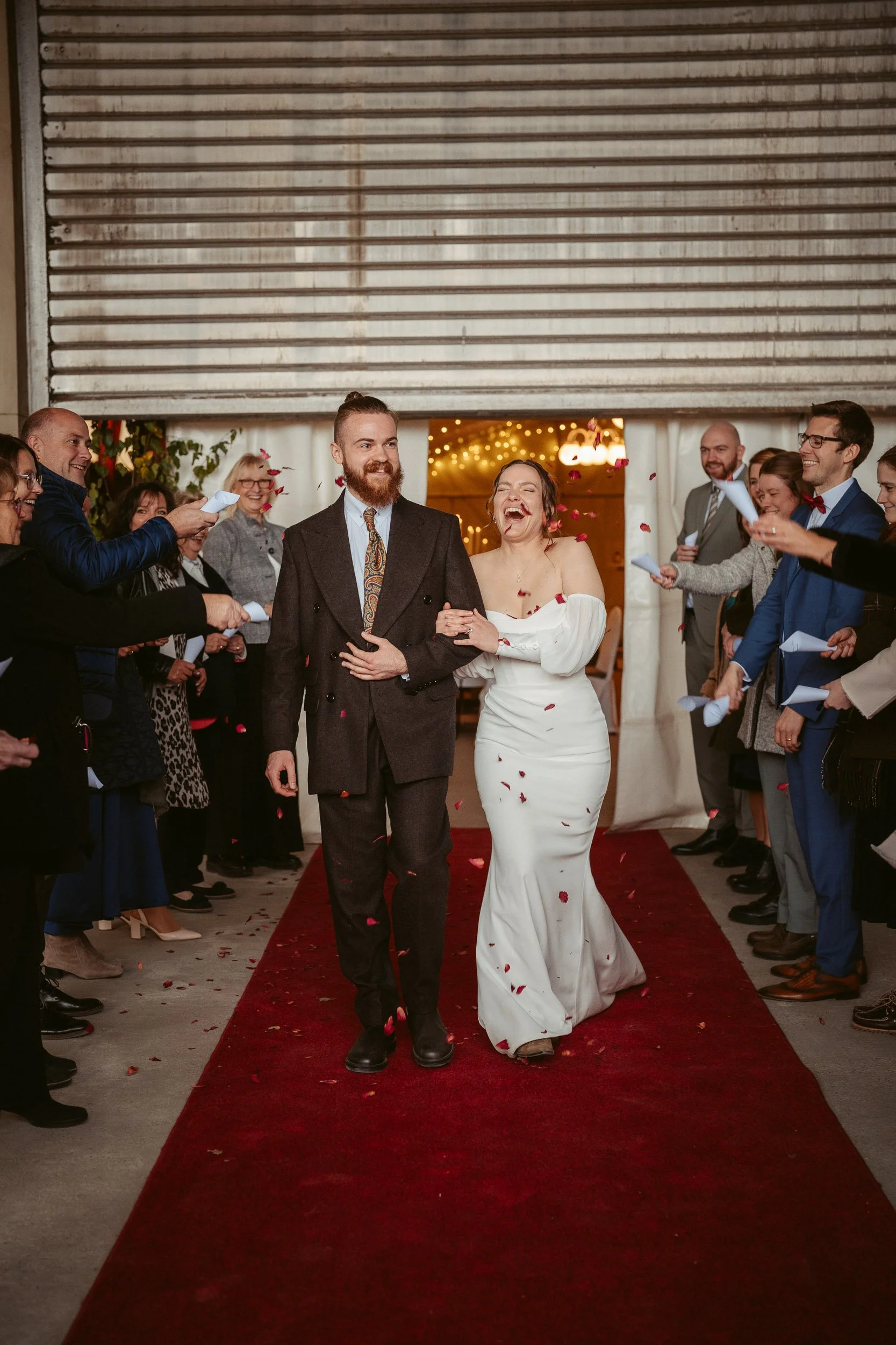 Ein Hochzeitsfoto mit einem lachenden Brautpaar, das Hand in Hand auf einem roten Teppich geht, umgeben von Gästen, die Konfetti werfen.