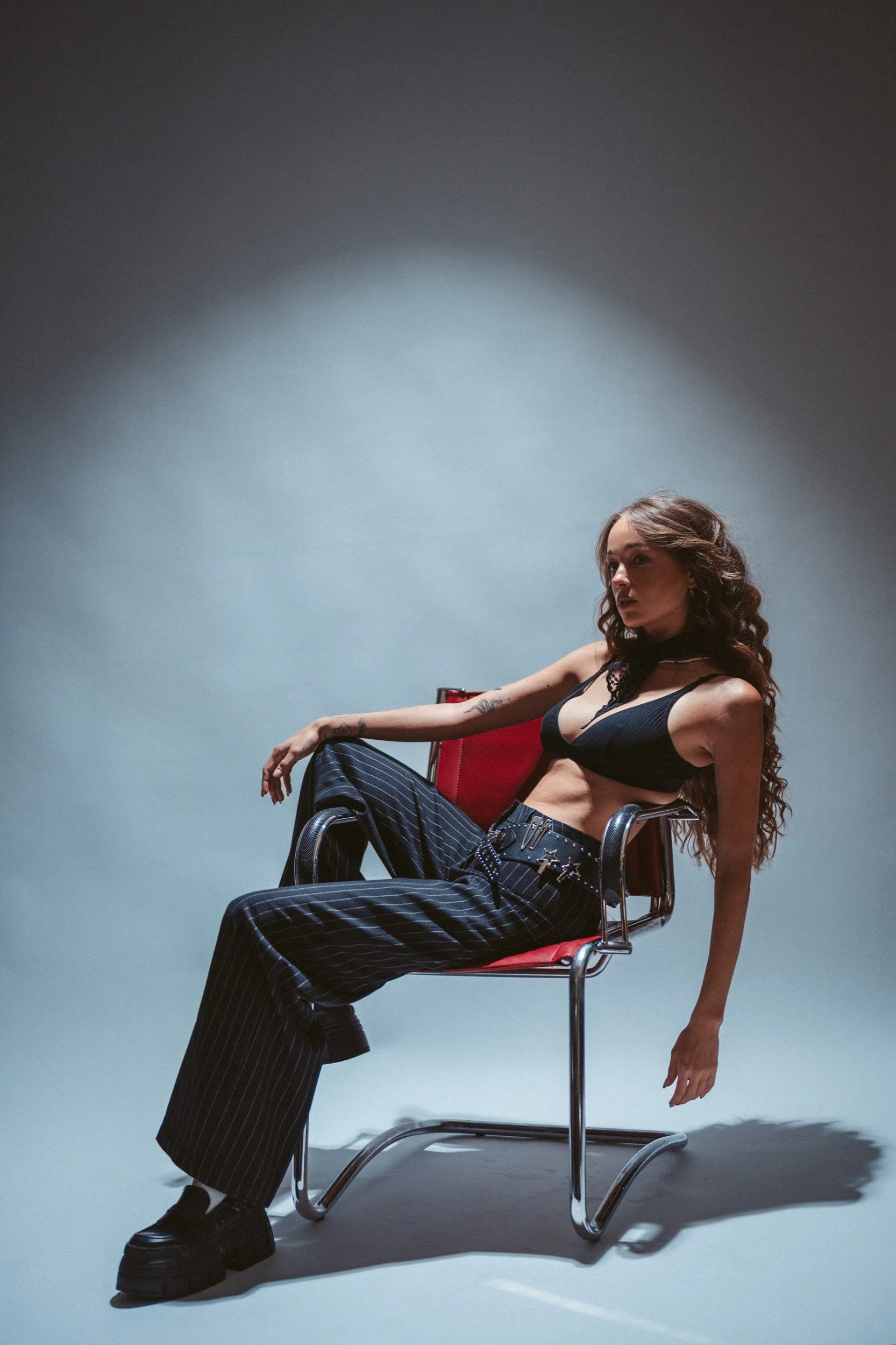 Frau mit langen, lockigen Haaren sitzt auf einem roten Stuhl, trägt eine schwarze Daniel-Kleid und schwarze, gestreifte Hosen, mit einem dunklen Hintergrund.