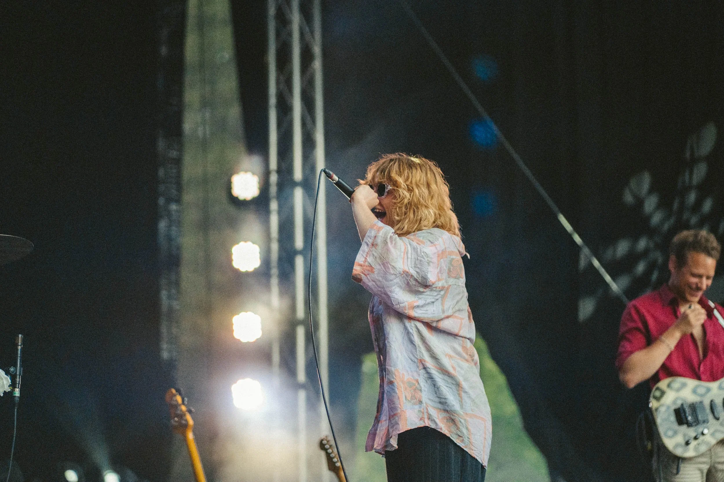 Eine weibliche Sängerin mit roten, lockigen Haaren trägt eine lockere, bunte Bluse und singt auf einer Bühne bei einem Konzert, im Hintergrund sind Musiker und Bühnenlichter sichtbar.
