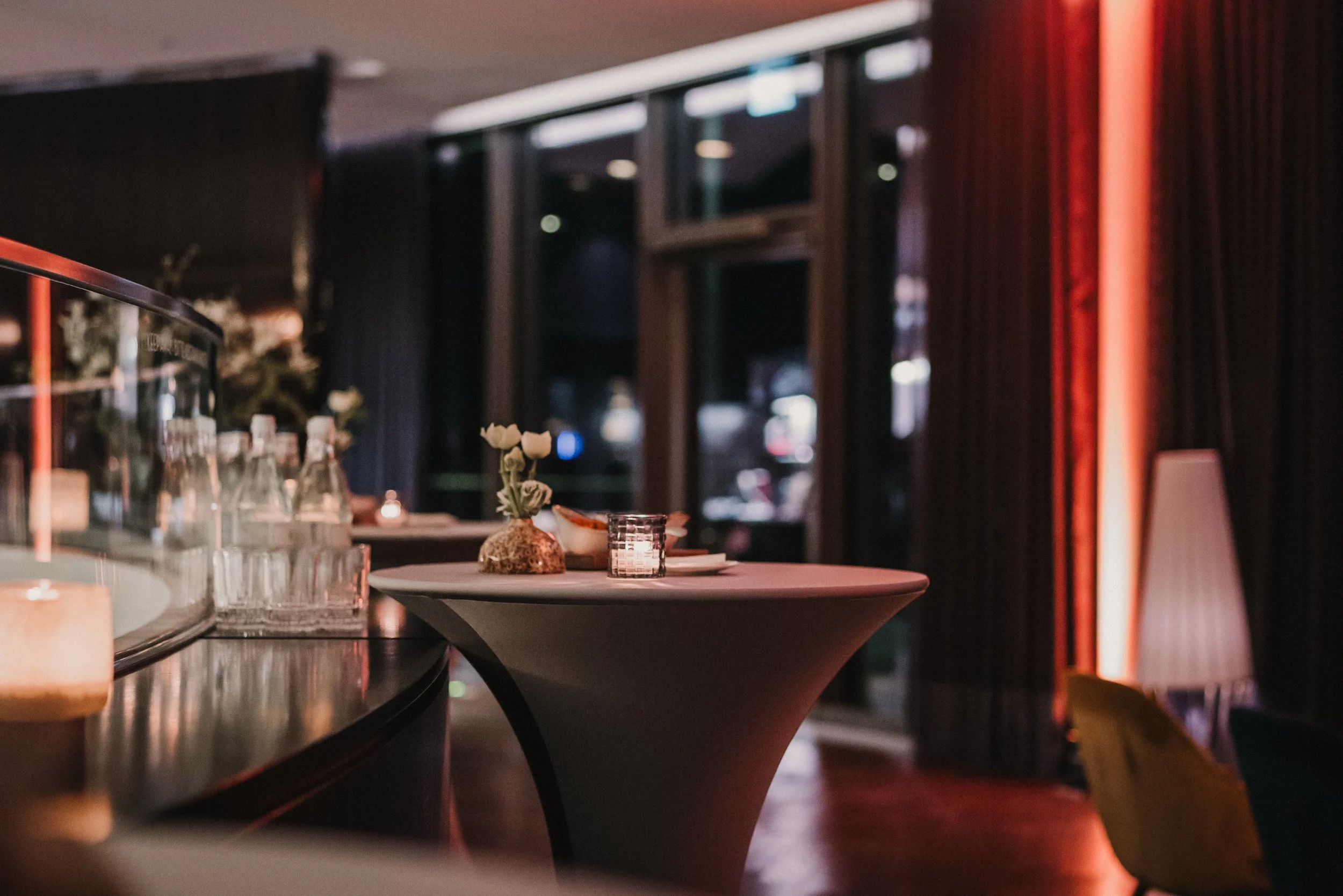 Innenraum eines eleganten Restaurants mit einem hohen, runden Tisch, weißen Blumen in einer Vase, Kerzenlichter und dunkler Dekoration, bei gedämpftem Licht.