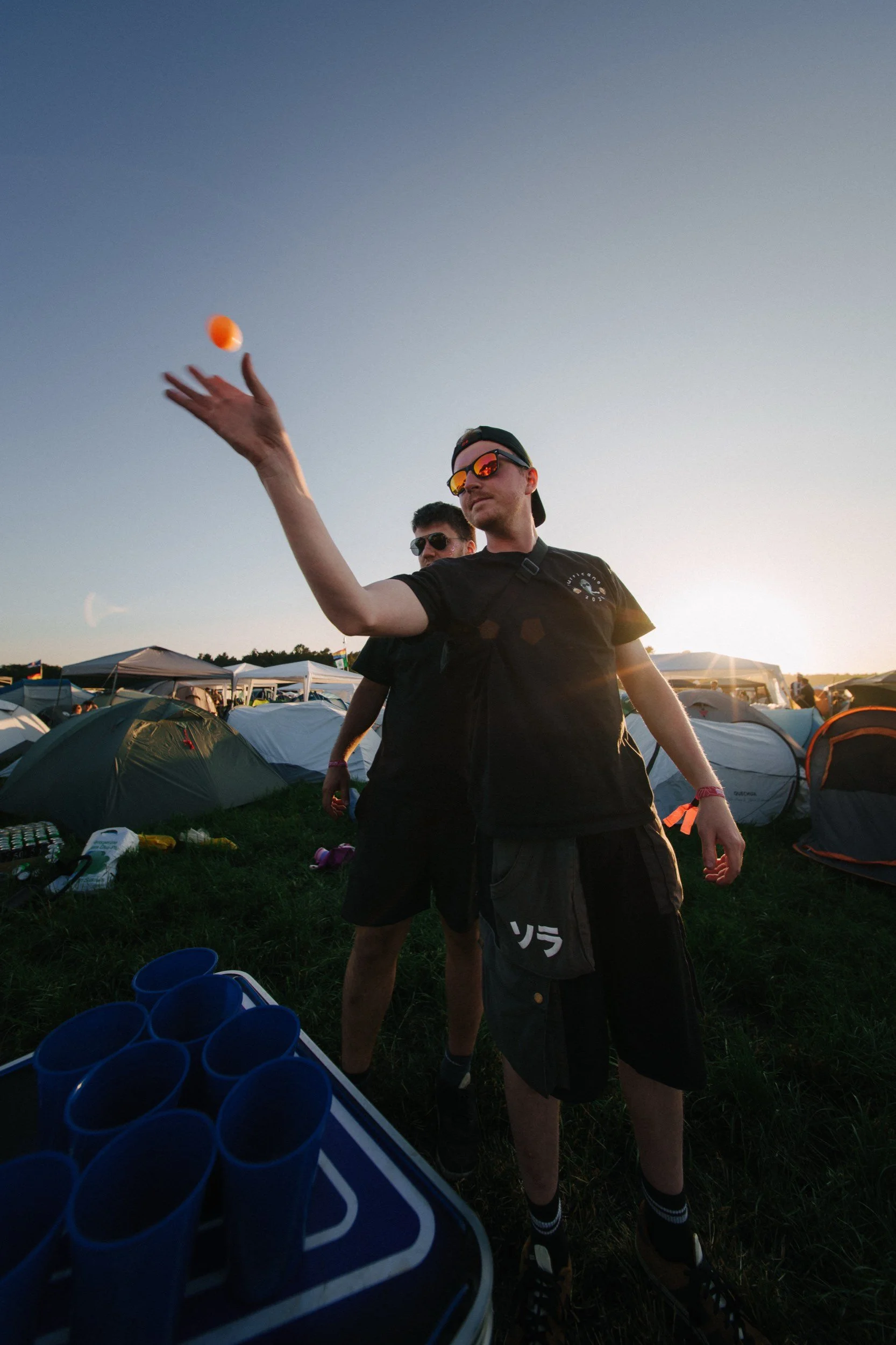 Zwei junge Männer auf einem Campingplatz beim Feiern, einer wirft einen orangefarbenen Ball in die Luft bei Sonnenuntergang, umgeben von Zelten.