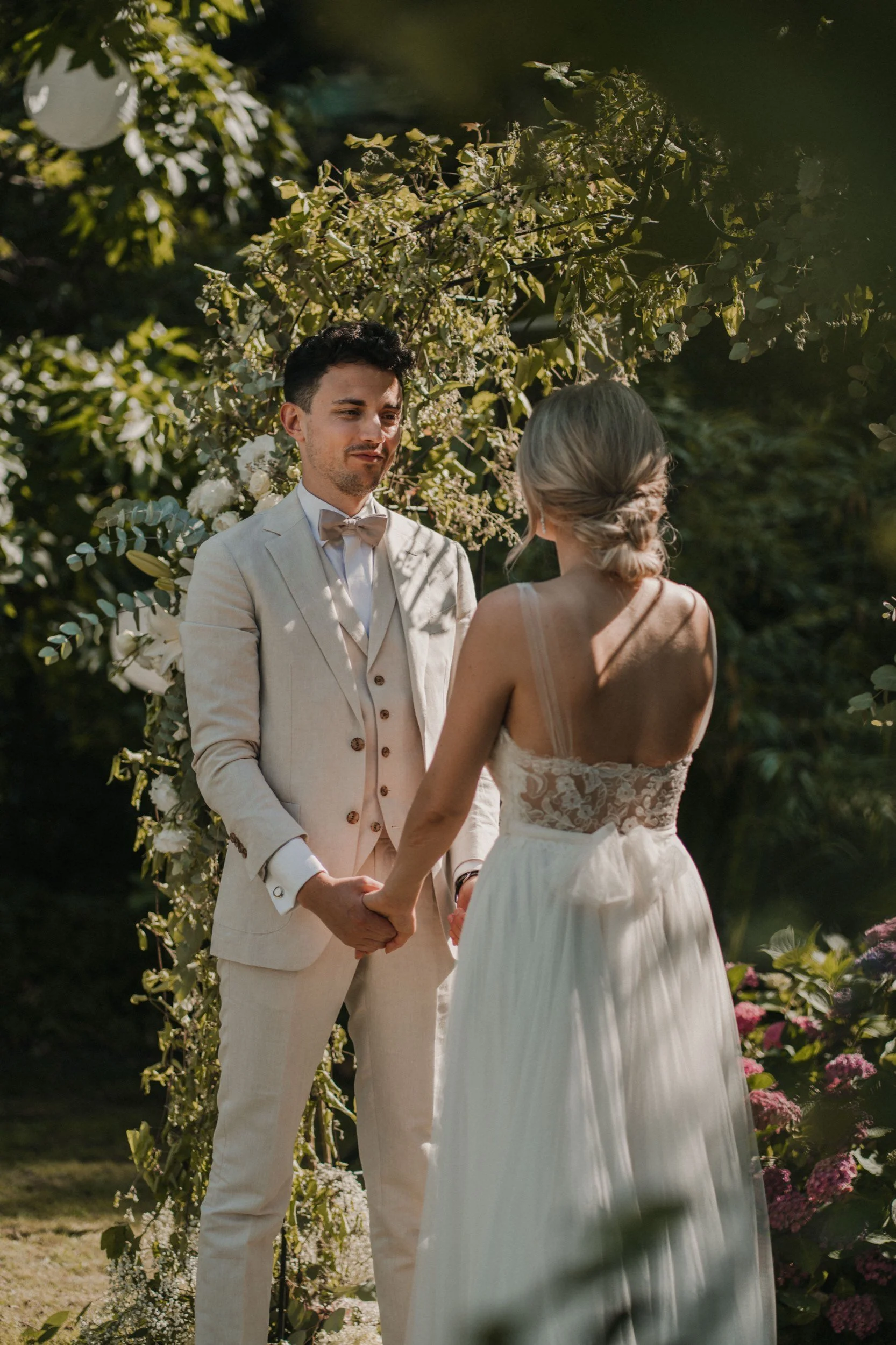 Ein Brautpaar bei einer standesamtlichen Hochzeit im Freien, die Hand haltend, vor einem Blumenbogen, umgeben von grünen Bäumen und bunten Blumen, bei Sonnenlicht.