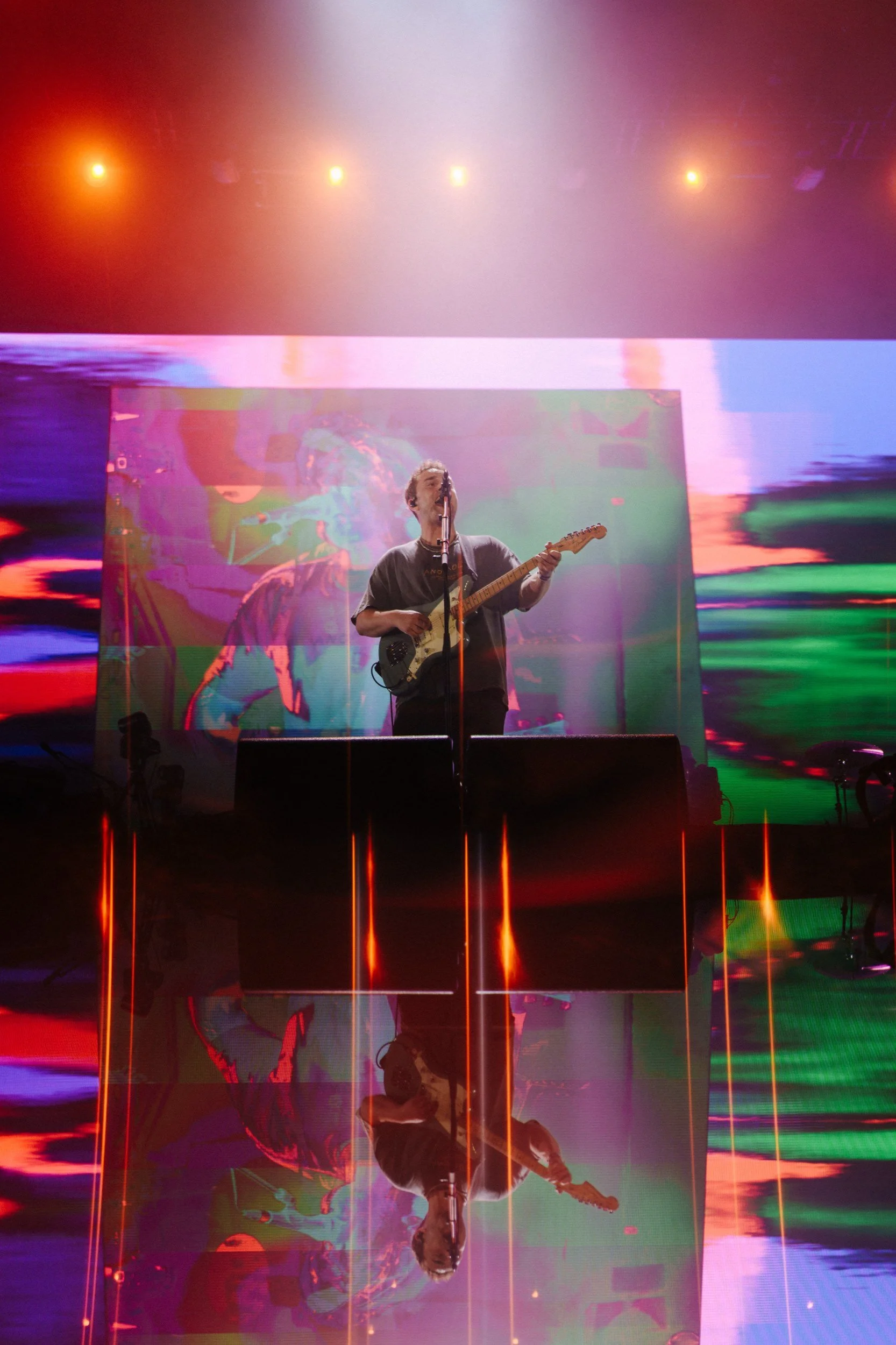 Ein Musiker spielt Gitarre auf einer Bühne mit bunten, abstrakten Lichteffekten und einem großen Bildschirm im Hintergrund.