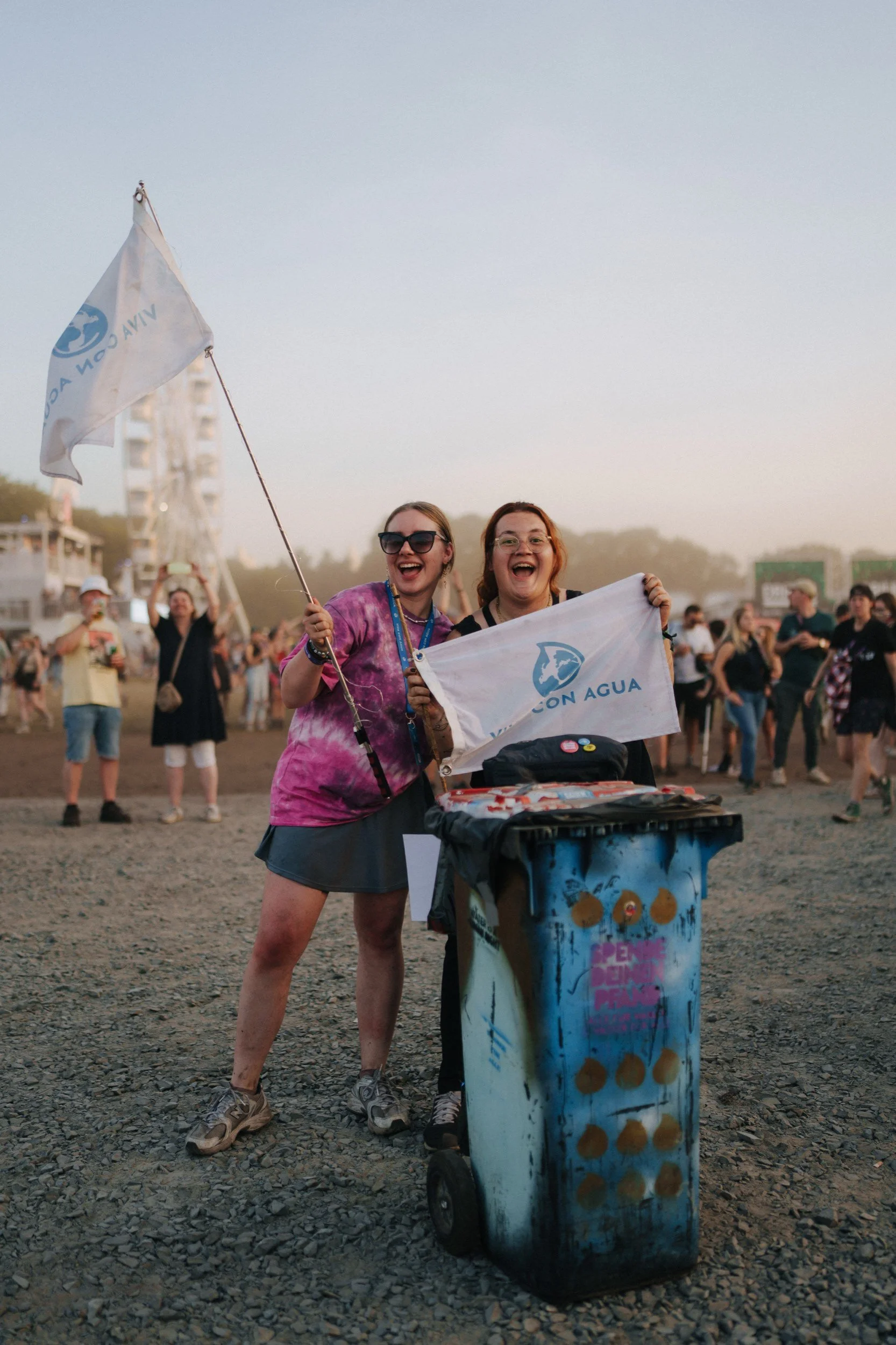 Zwei lachende Frauen, die Flaggen mit der Aufschrift 'VIVA CON AGUA' halten, im Hintergrund viele Menschen bei einem Festival auf einem offenen Feld.