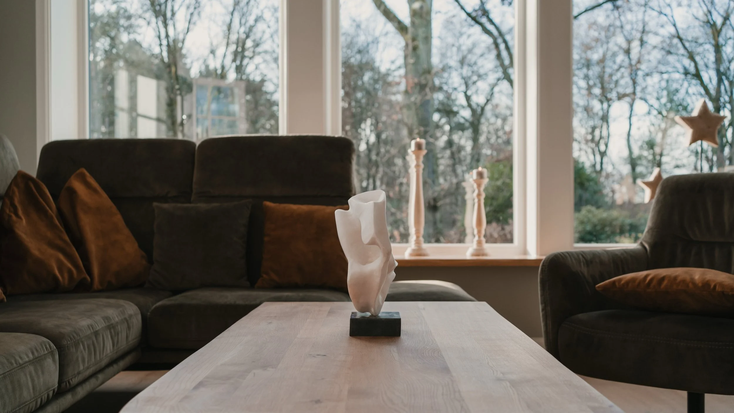 Wohnzimmer mit braunen Sofas, dunkler Samt, braunen Kissen, einem weißen Skulptur auf einem Holztisch, zwei Kerzenhalter und Fenster mit Blick auf Bäume im Garten.