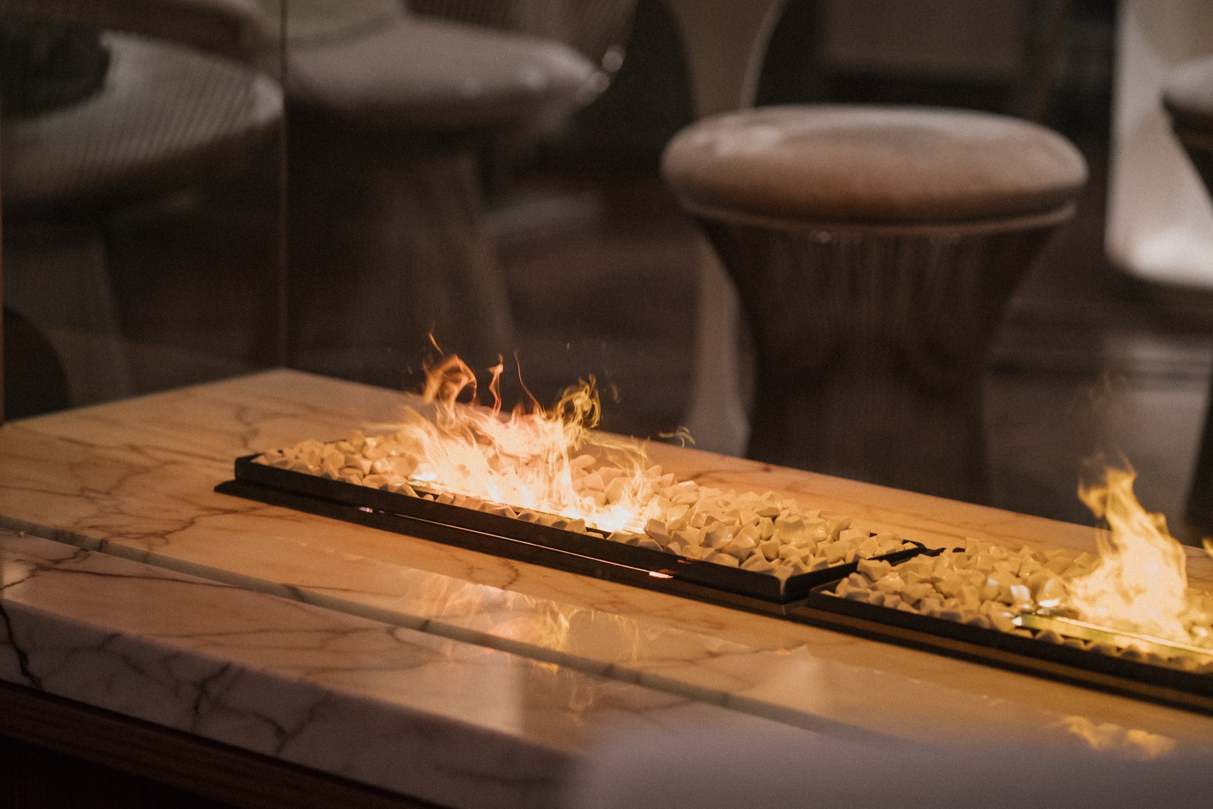 Ein Tisch mit Feuer und Kerzen in Form von weißen Steinen auf einer Marmorplatte, im Hintergrund mehrere Stühle.