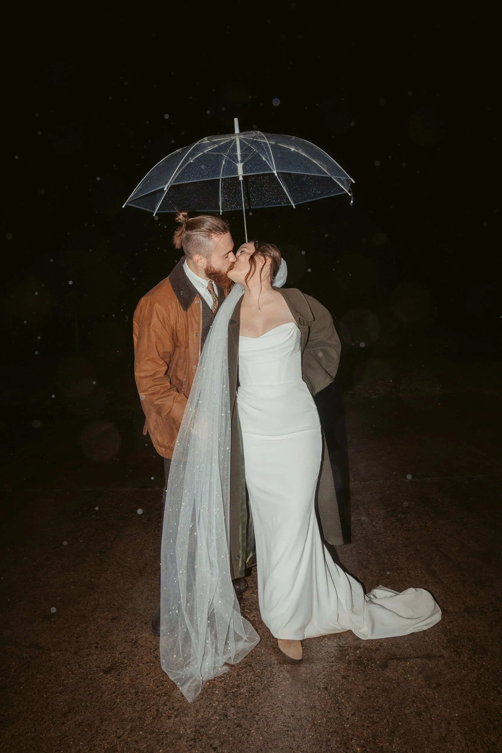 Ein verliebtes Paar in Hochzeitskleidung küsst sich unter einem transparenten Regenschirm bei Regen in der Nacht.