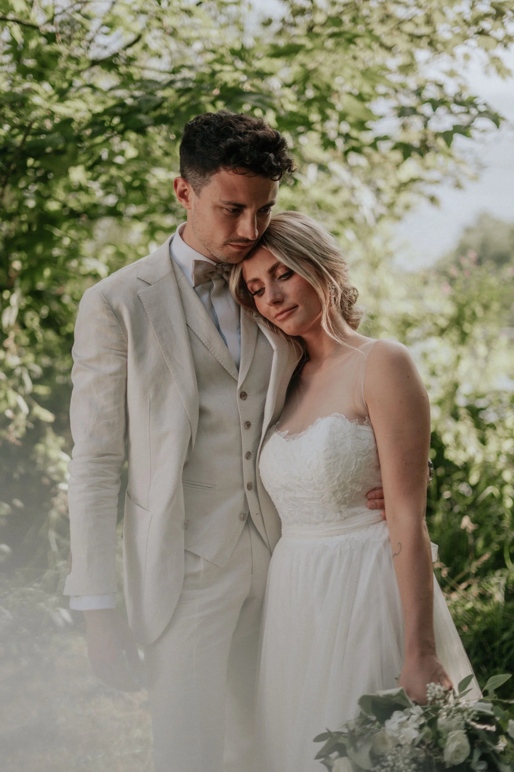 Ein Brautpaar umarmt sich im Freien, wahrscheinlich bei einer Hochzeit, beide tragen formelle weiße Kleidung, umgeben von grünen Bäumen.