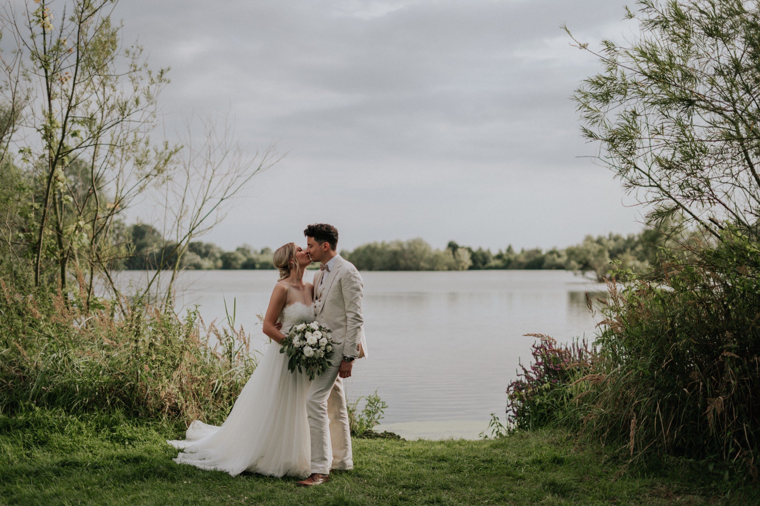 Ein Brautpaar bei ihrer Hochzeit am See, umgeben von Bäumen, es regnet leicht.