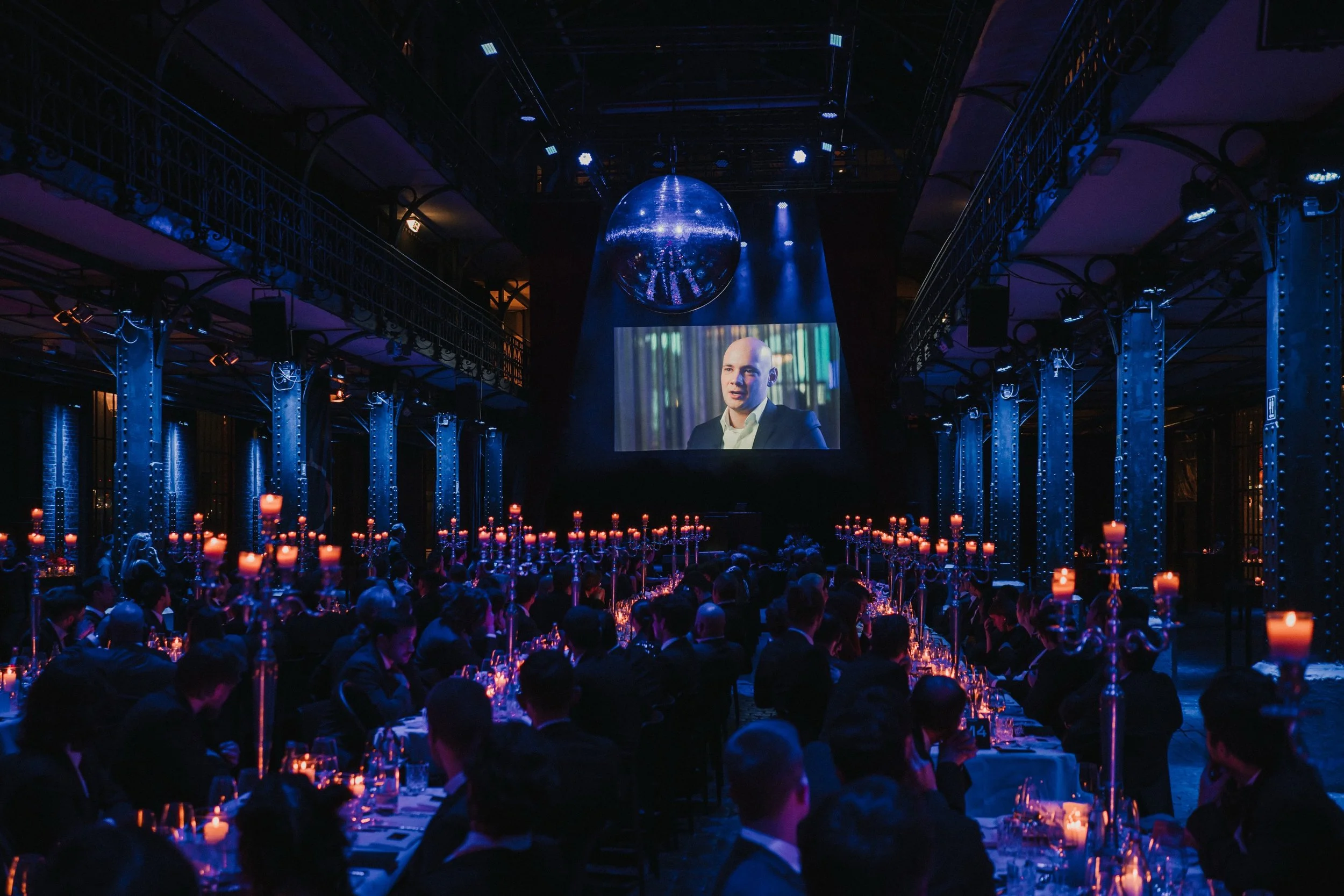 Ein dunkler Veranstaltungsraum mit langen gedeckten Tischen, Kerzen und Menschen in dunkler Kleidung, die einem Film- oder Präsentationsereignis zuschauen, mit einer großen Leinwand, auf der ein Mann in einem Anzug erscheint, und einem Disco-Kugel üb