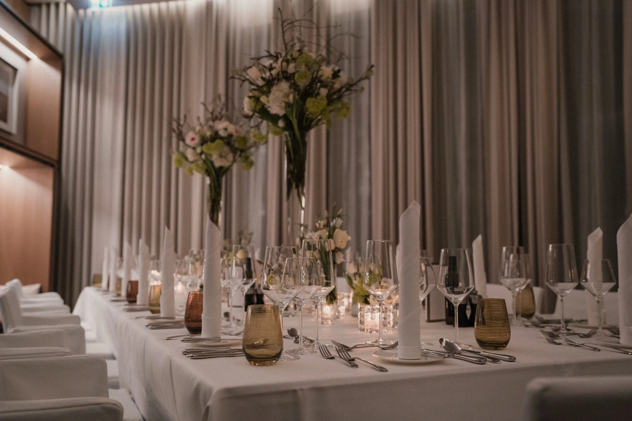 Eleganter gedeckter Esszimmertisch mit weißen Tischdecken, hohen weißen Kerzen, Weingläsern, kleinen Kerzenleuchtern und Blumenarrangements in Vasen, vor langen grauen Vorhängen.