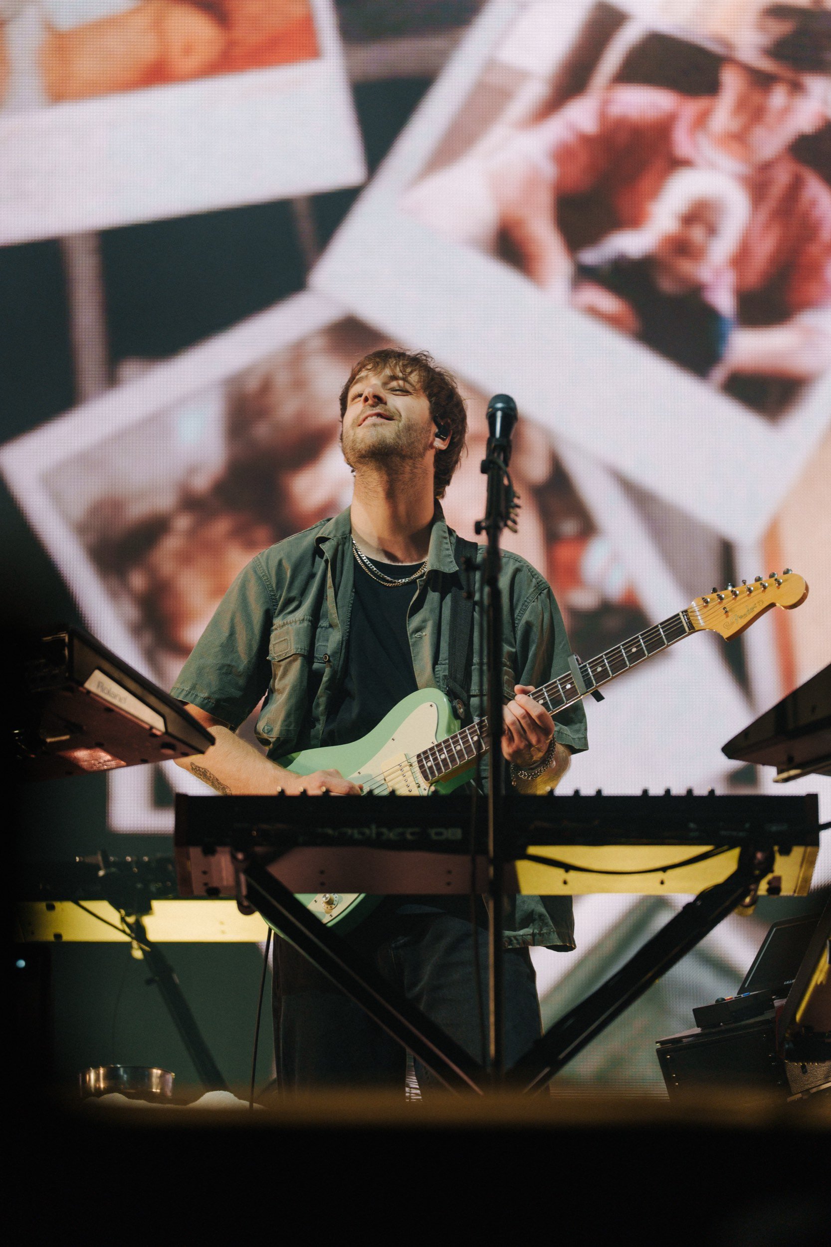 Ein Musiker mit einem E-Gitarrenspiel auf der Bühne, im Hintergrund eine große Leinwand mit Fotos von Menschen.